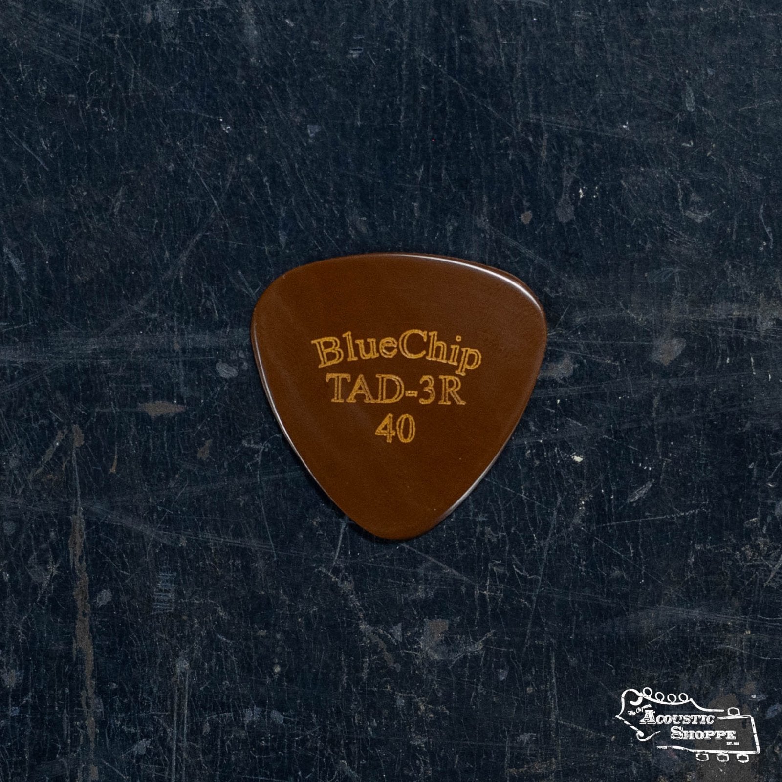 A brown BlueChip Picks (TAD40-3R) pick with BlueChip TAD-3R 40 in gold text rests on a dark, scratched surface. The Acoustic Shoppe logo appears at the bottom right, highlighting this premium BlueChip acoustic accessory.