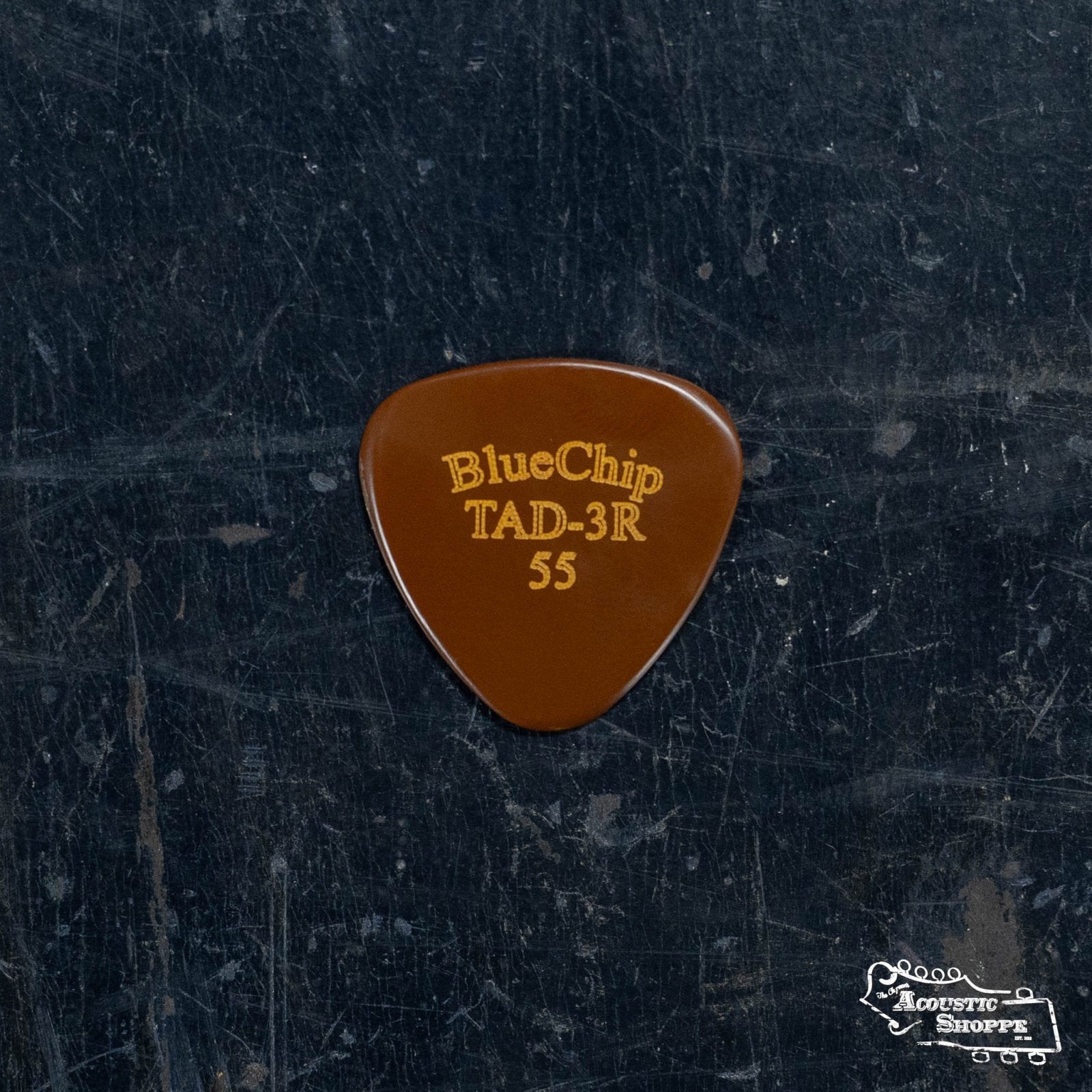 A BlueChip Pick (TAD55-3R) by BlueChip, brown with rounded edges and gold lettering, displays a small Acoustic Shoppe logo in the bottom right corner as it rests on a dark, textured surface.