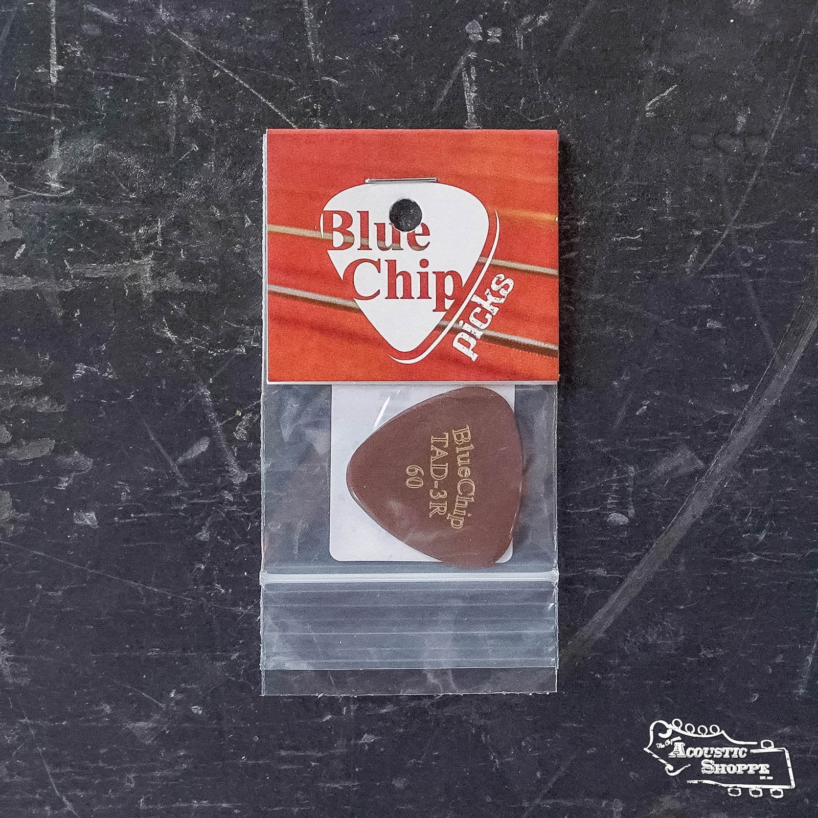 A BlueChip Picks (TAD60-3R), hand beveled for precision, rests in a small plastic bag with a red and white BlueChip label, photographed on a dark textured surface. The Acoustic Shoppe logo appears in the bottom right corner.
