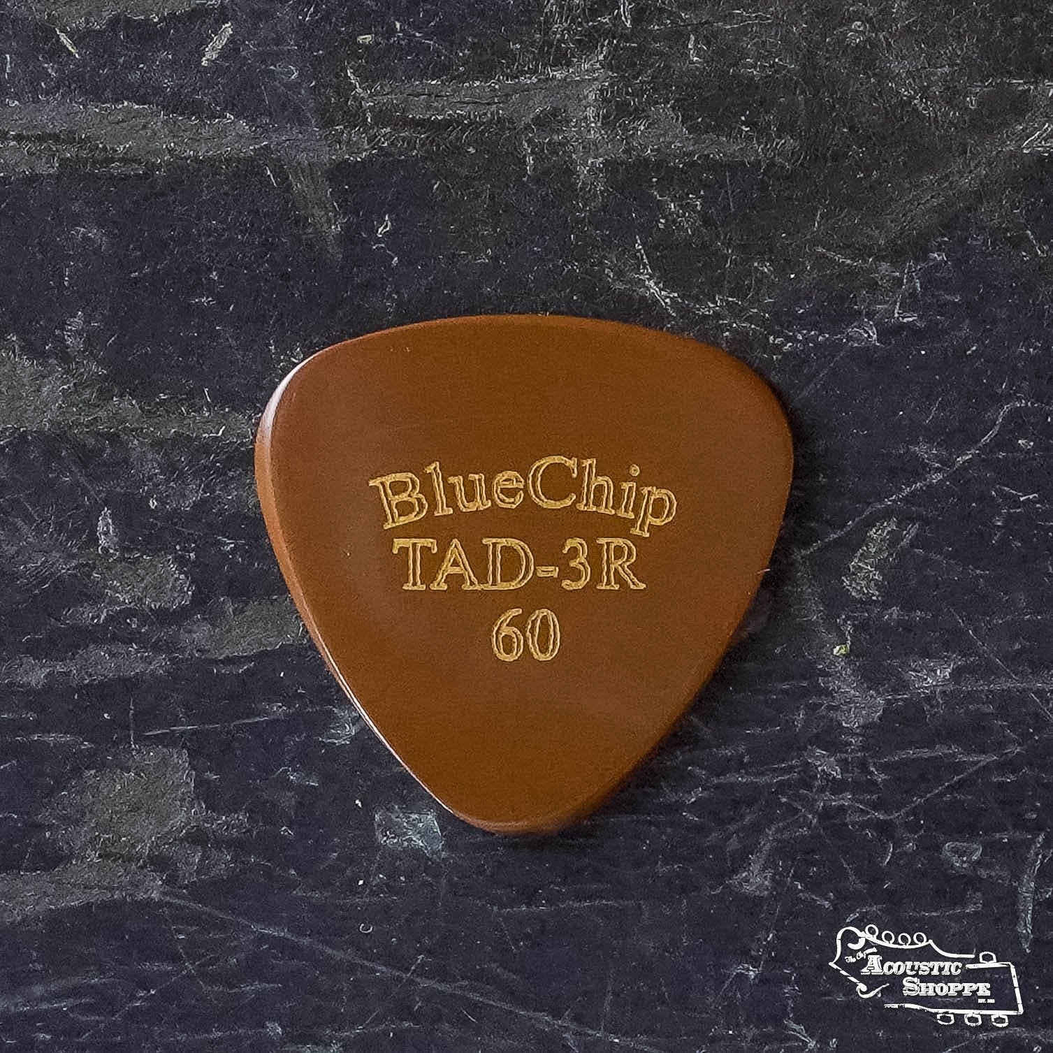 A BlueChip Picks (TAD60-3R) hand-beveled brown guitar pick by BlueChip rests on a dark textured surface, with The Acoustic Shoppe logo in the lower right corner.