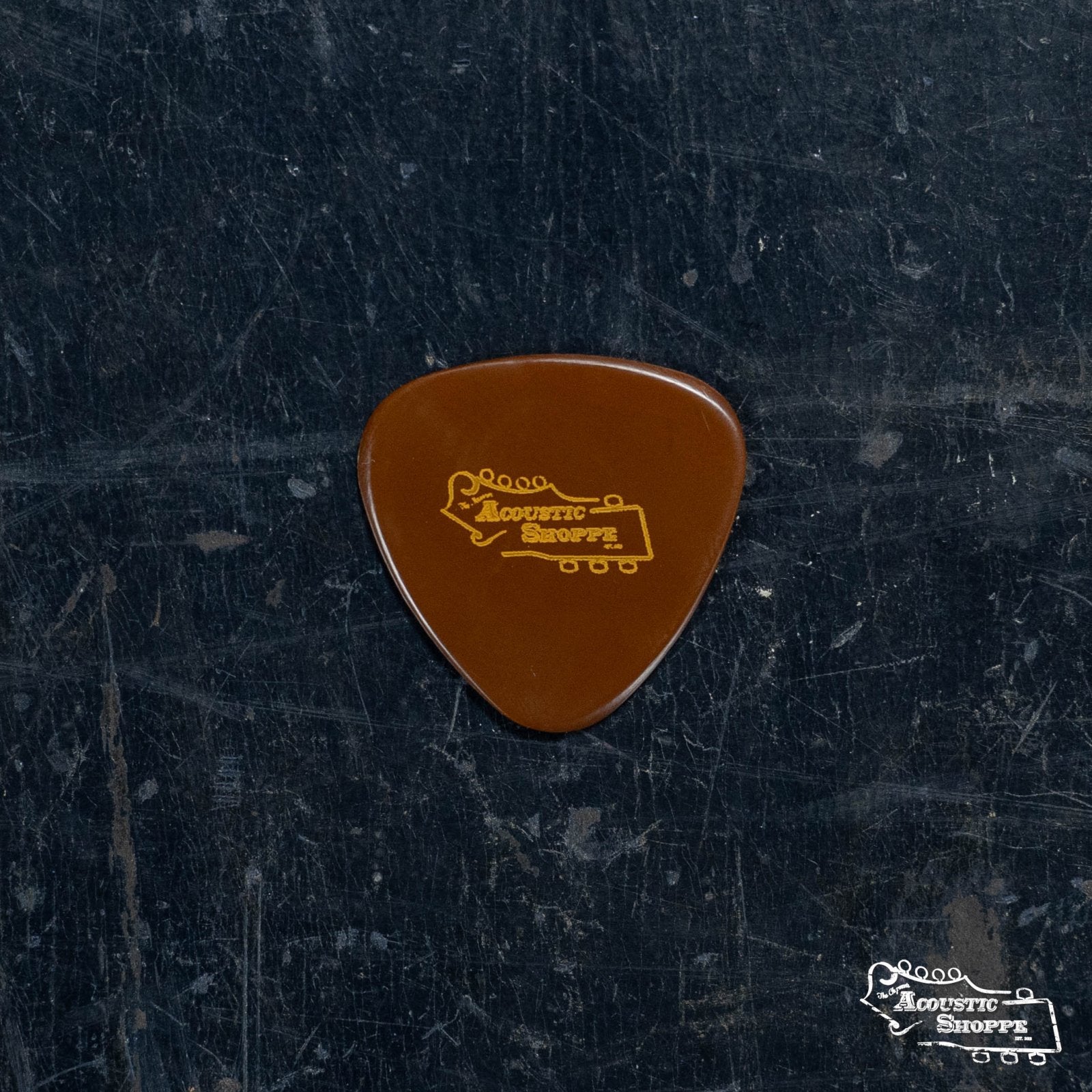 A BlueChip Picks (TAD40-3R) guitar pick by BlueChip, with the Acoustic Shoppe and guitar headstock logo in yellow, shown on a dark, scratched surface—ideal for acoustic music enthusiasts.