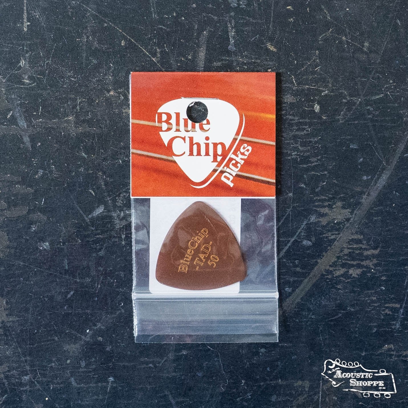 A BlueChip Picks (TAD50) guitar pick by BlueChip, showing gold lettering and packed in branded red and white packaging with a small plastic bag, rests on a dark, worn surface—perfect for acoustic accessories enthusiasts.