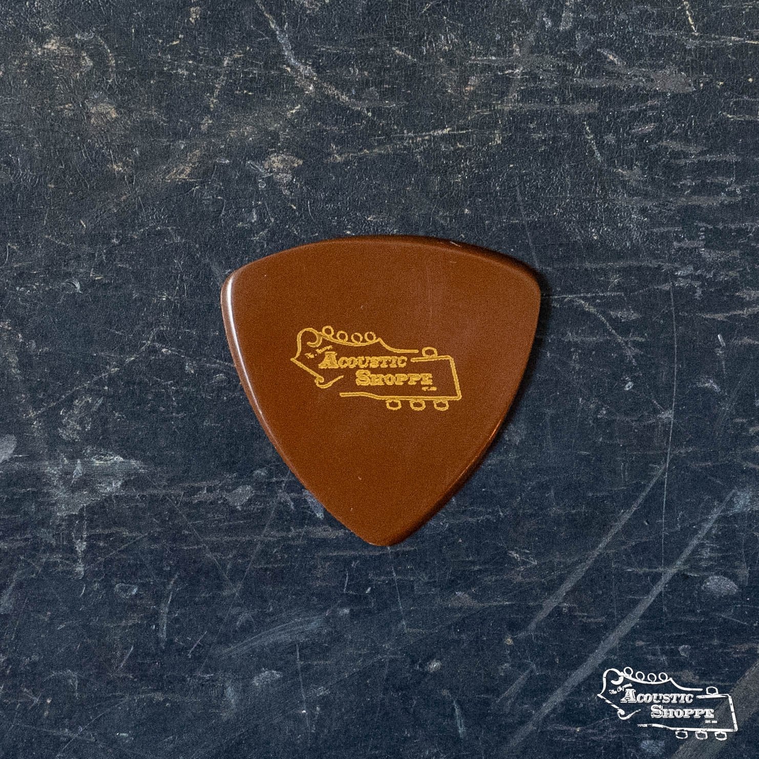A BlueChip Picks (TAD50) by BlueChip, featuring Acoustic Shoppe and a guitar headstock design in yellow on a brown pick, rests on a dark textured surface—an ideal addition to your acoustic accessories collection.