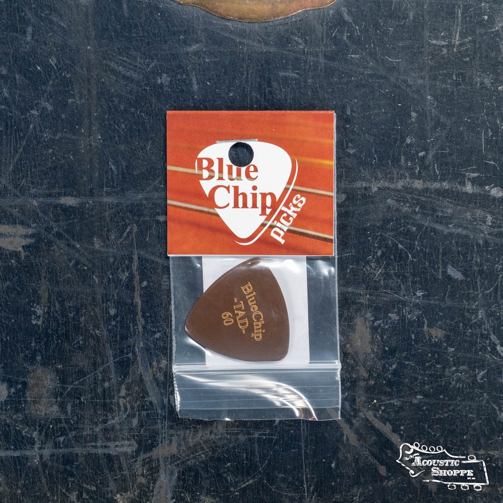 A BlueChip Picks (TAD60) guitar pick by BlueChip, packaged with an orange and white card, rests on a scratched dark metallic surface featuring “The Acoustic Shoppe” logo—ideal for boutique guitar accessory enthusiasts.
