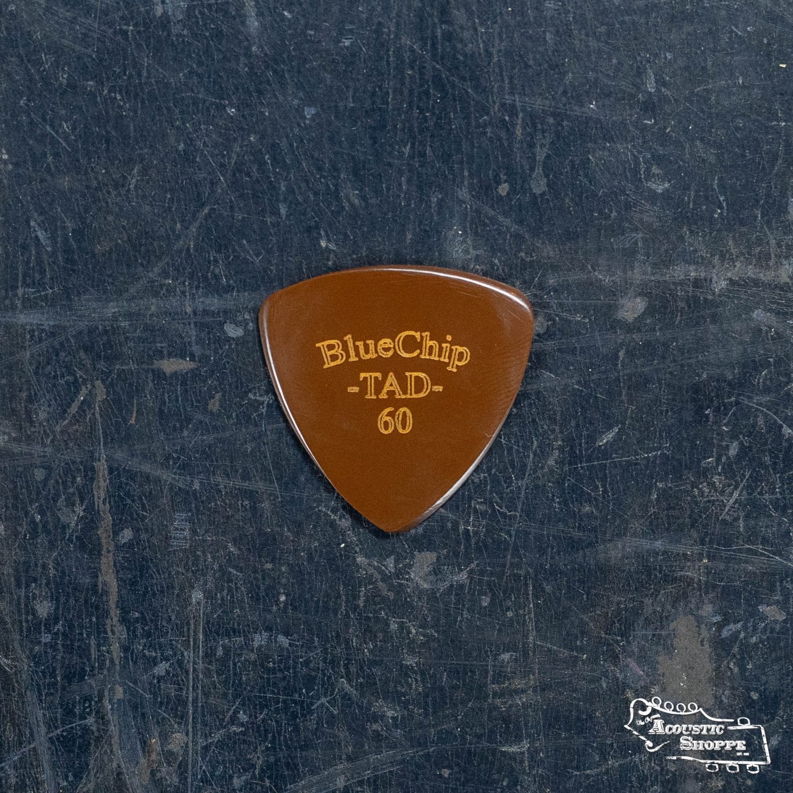 A BlueChip Picks (TAD60) guitar pick from BlueChip, renowned for acoustic excellence, is placed on a dark, textured surface with faint scratches. The Acoustic Shoppe logo is visible in the lower right corner.