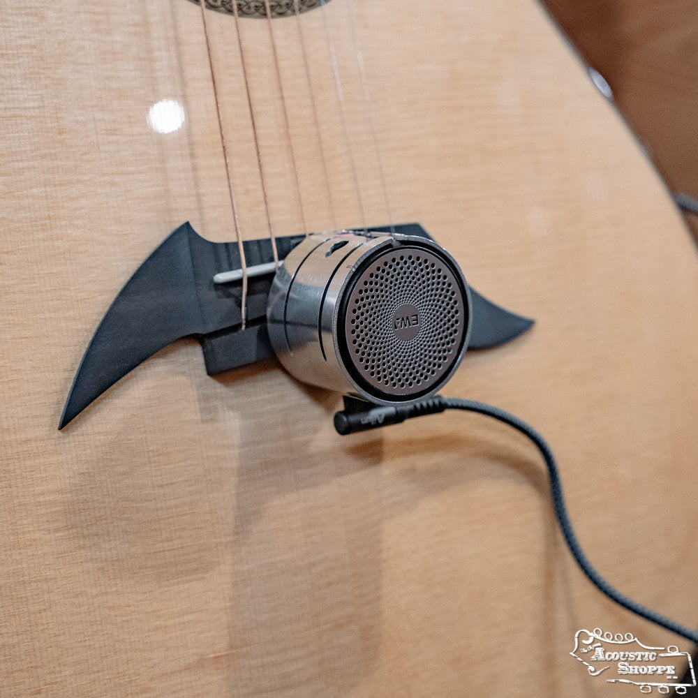 A close-up of a guitar with an EM3 microphone attached near the soundhole, featuring a black bat-shaped pickguard. The mic is connected via cable to the DrHerringBone Tone Traveler Trio Package acoustic enhancement system.