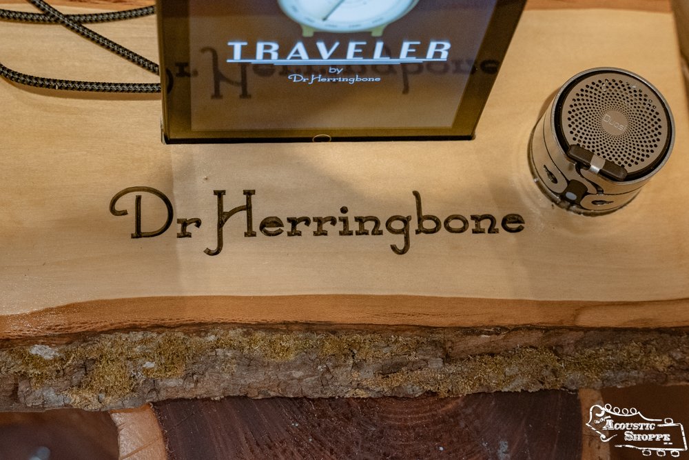 A wooden display engraved with DrHerringBone holds a small speaker clamp and sign for the Dr. Herringbone Tone Traveler Trio Package, an acoustic instrument enhancement product. The Acoustic Shoppe logo is in the bottom right corner.