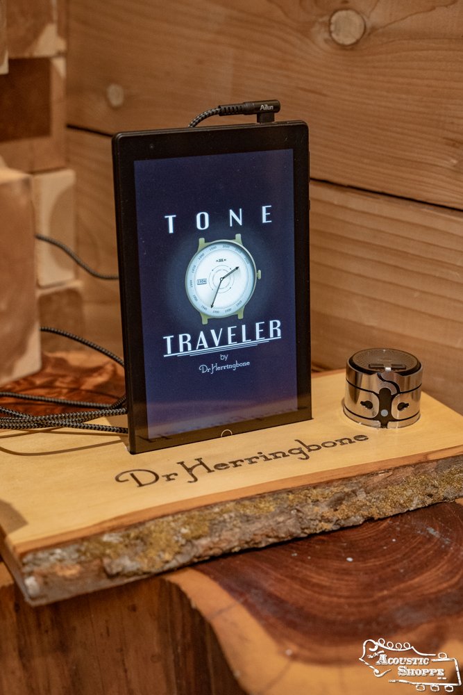 A tablet displays the Dr. Herringbone Tone Traveler Trio Package by DrHerringBone on a wood slab engraved with the logo. Nearby, a coiled cable and speaker clamp suggest acoustic instrument enhancement amid rich wood textures.