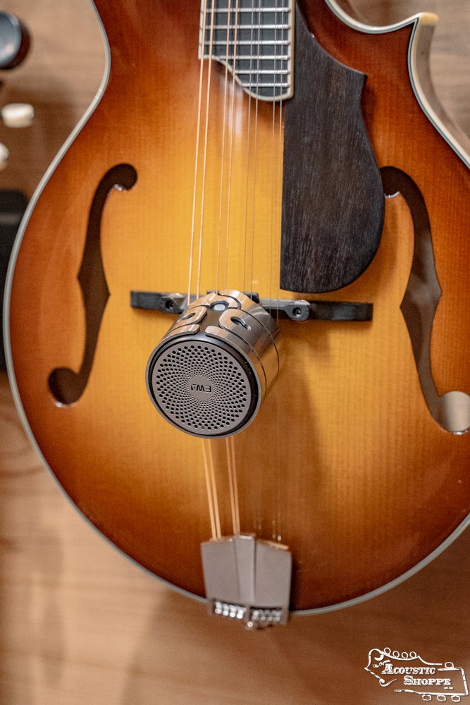 A close-up of the DrHerringBone Tone Traveler Trio Package shows its speaker clamp near the sound hole of a sunburst mandolin, demonstrating how this accessory enhances acoustic tone by working directly with the instrument’s body and strings.