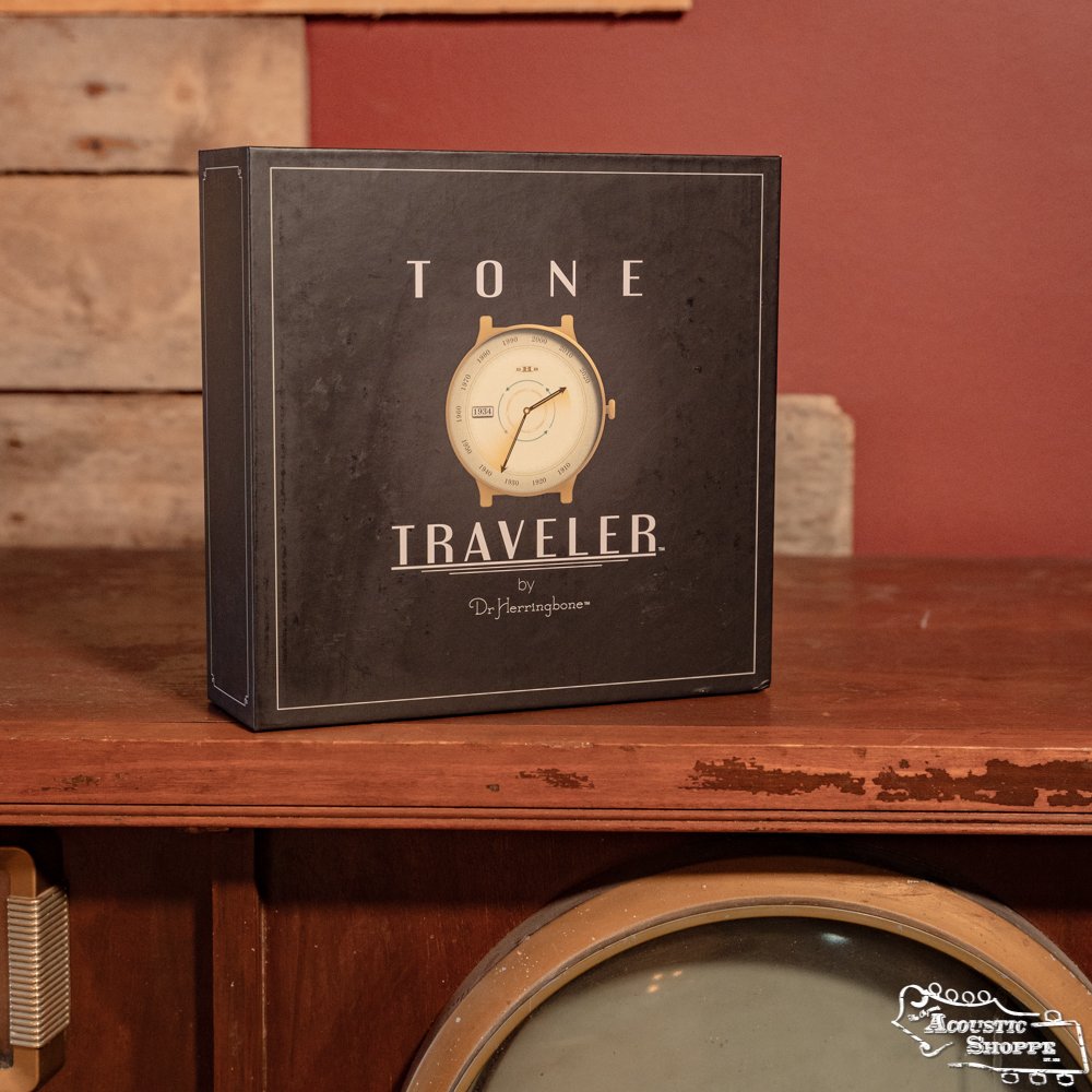 A black square box labeled Dr. Herringbone Tone Traveler Trio Package by DrHerringBone sits on a wooden cabinet in a rustic room with a red wall, featuring a clock face graphic and designed to enhance acoustic instruments.