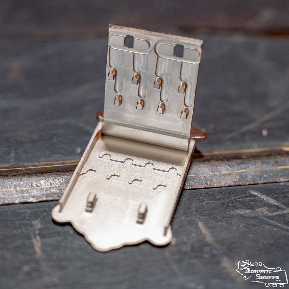 A close-up of the Axiom Inc. James Nickel Traditional Mandolin Tailpiece with an open lid, showing slots for strings. The tailpiece, inspired by classic mandolin accessories, rests on a worn, dark wooden surface.