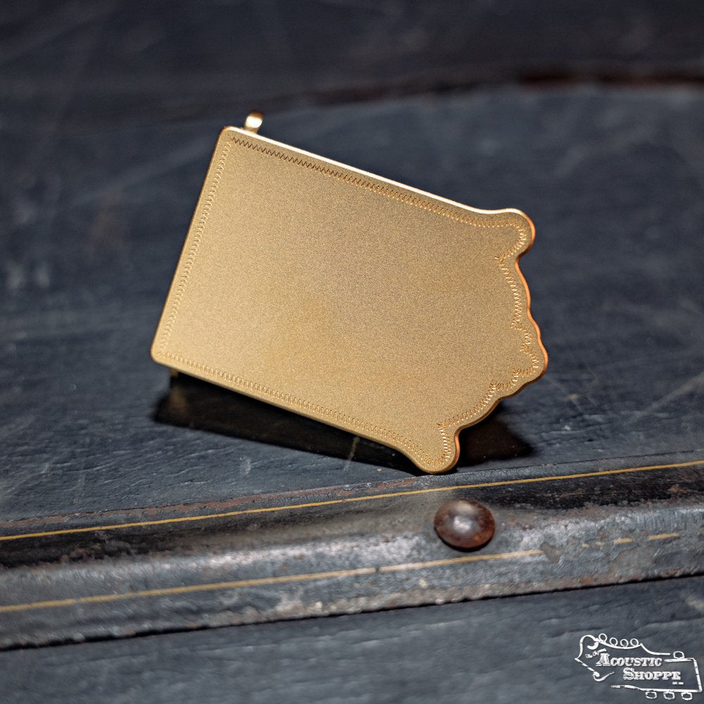 The James Gold Traditional Mandolin Tailpiece by Axiom Inc.—a gold-colored, Iowa-shaped metal piece—rests on a dark textured surface near a metal rivet.