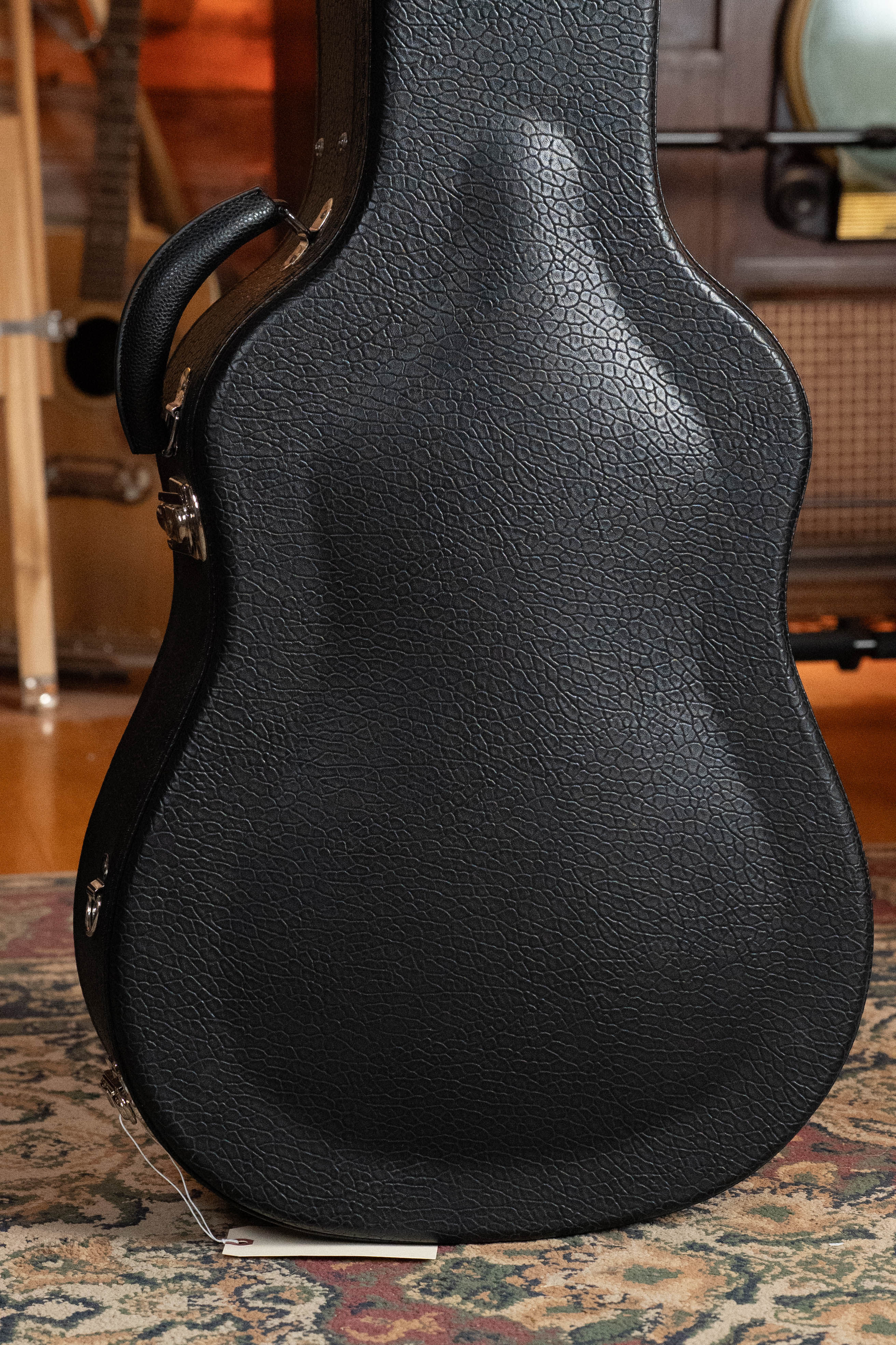 A black textured guitar case for the Bourgeois TAS LSH Professional Series Satin Dreadnought Guitar (*Pre-Order 25% Deposit ONLY*) stands upright on a patterned rug, featuring a side handle and white tag; wooden furniture and a vintage radio are in back.
