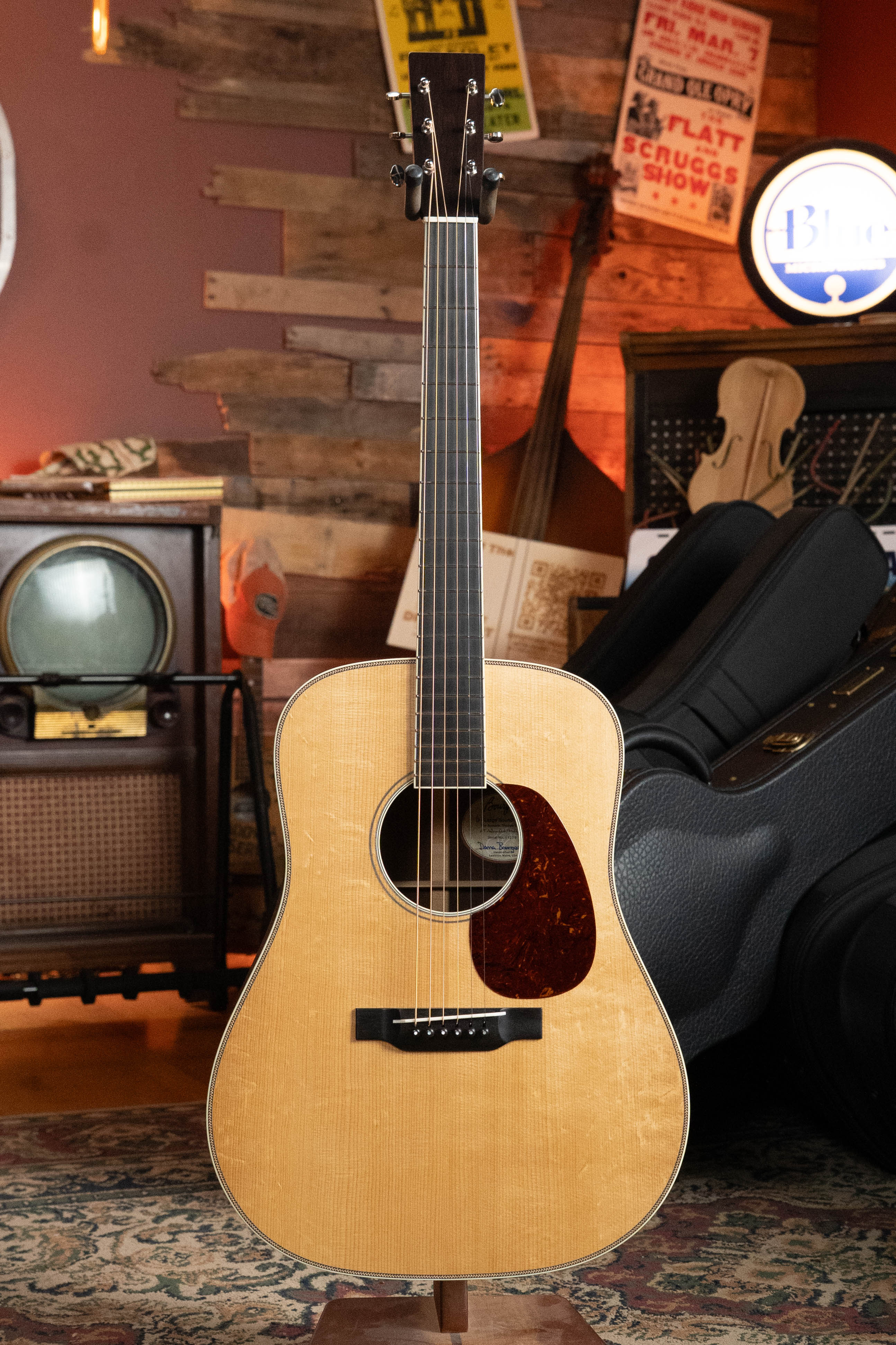 A *Pre-Order 25% Deposit ONLY* Bourgeois TAS LSH Professional Series Satin Dreadnought Guitar stands upright with its natural finish in a cozy vintage-style room adorned with musical instruments, posters, and retro decor.