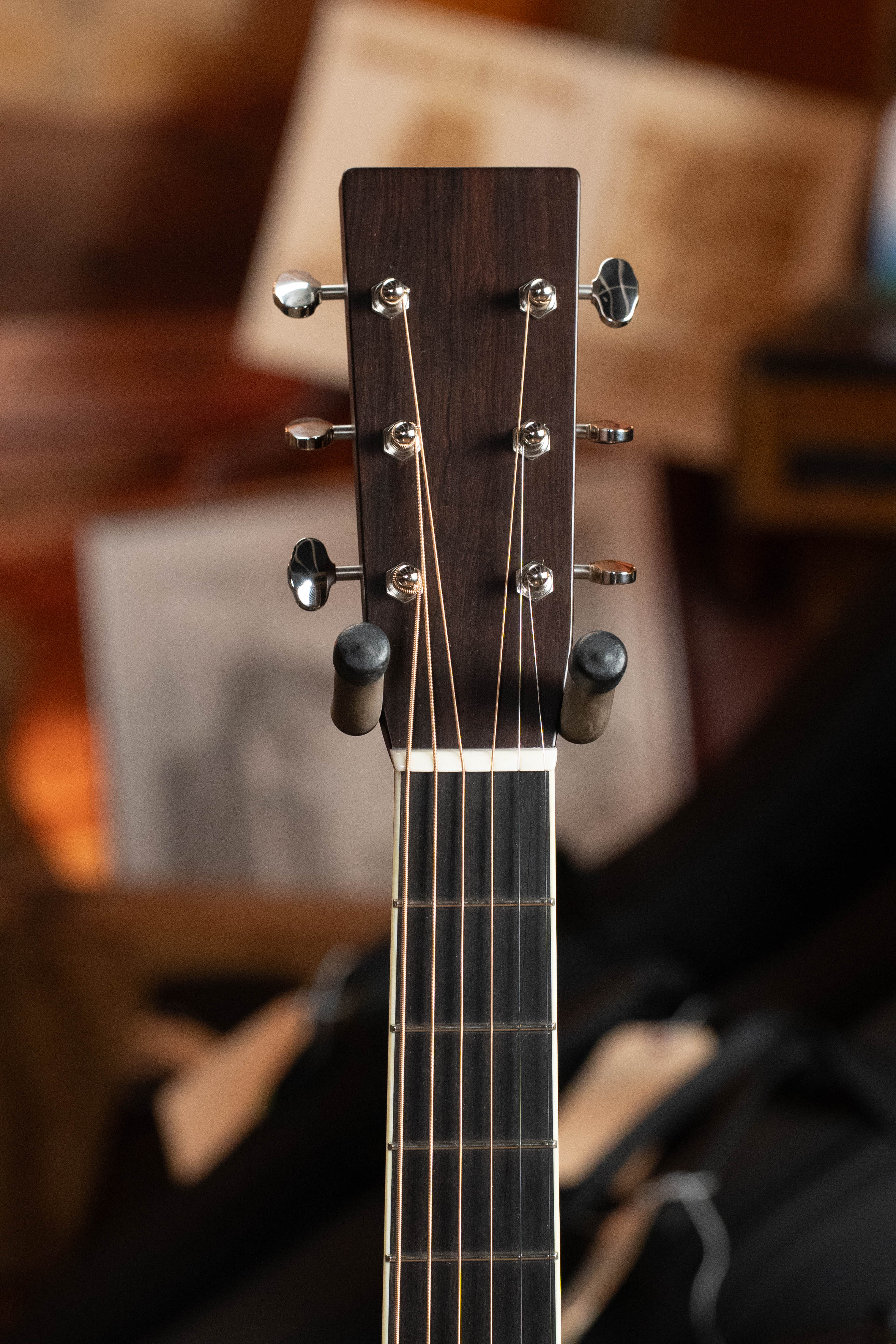 Close-up of a Bourgeois TAS LSH Professional Series Satin Dreadnought Guitar headstock, with six tuners, on a wall hanger; blurry background with indistinct papers and objects. *Pre-Order 25% Deposit ONLY.*.