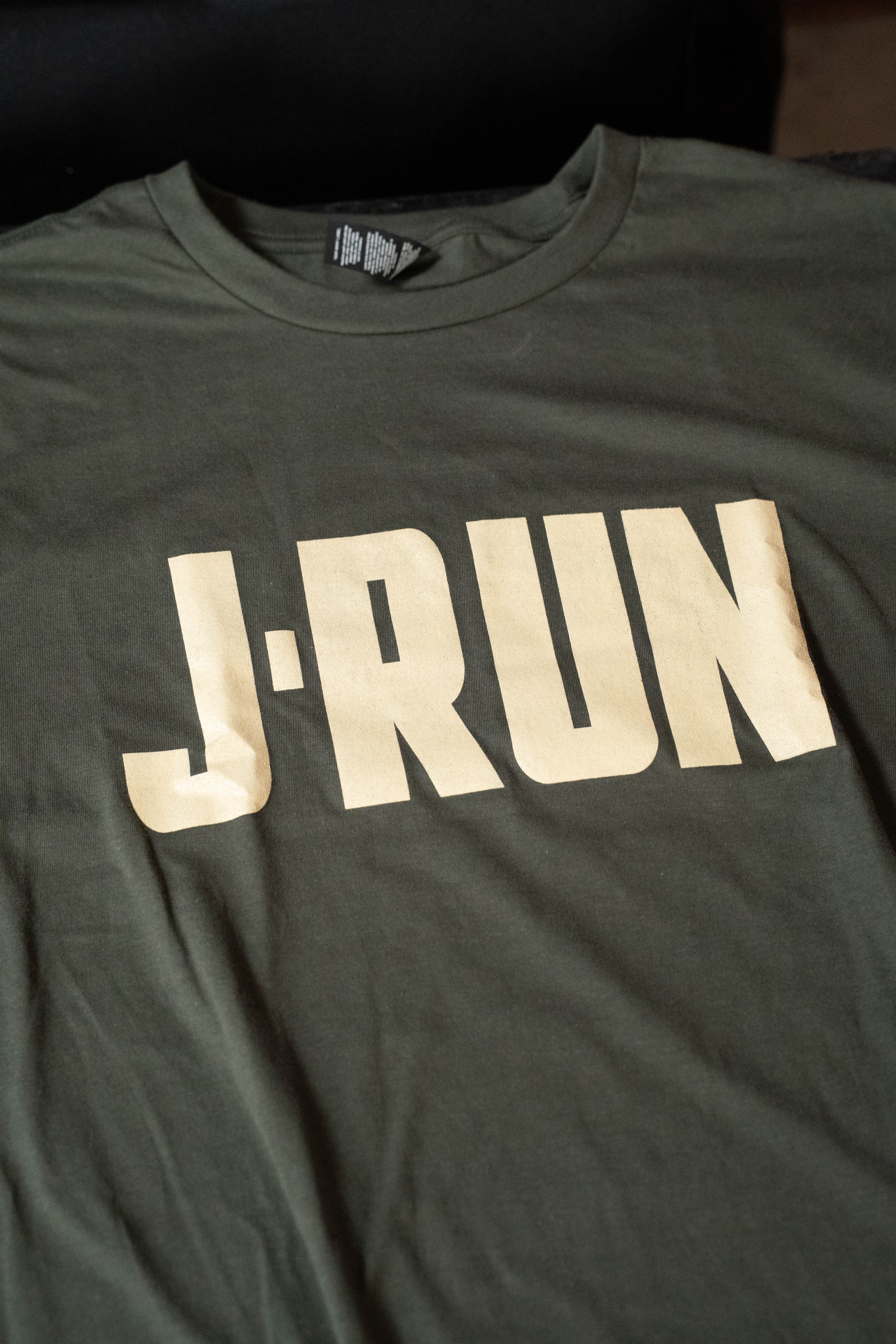 The TAS J-Run Olive Green Shirt w/Double Emblems by Amplify Screen Printing & Promotions features bold beige J-Run graphics—available now at Acoustic Shoppe.