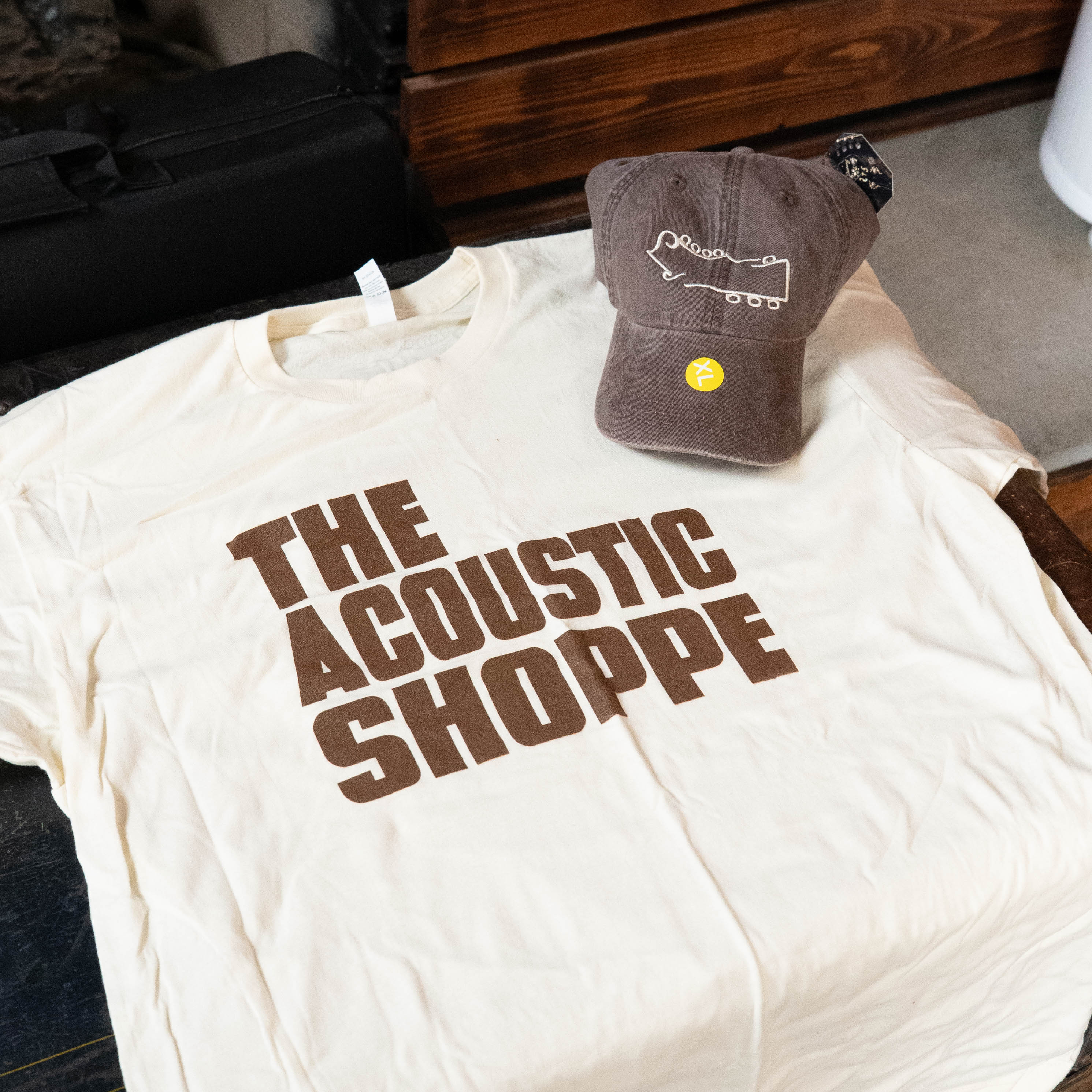 The TAS Logo Shirt and Hat Bundle by Amplify Screen Printing & Promotions features a cream shirt with bold brown THE ACOUSTIC SHOPPE lettering and a hat with a guitar outline and yellow smiley face, displayed on a wooden surface.