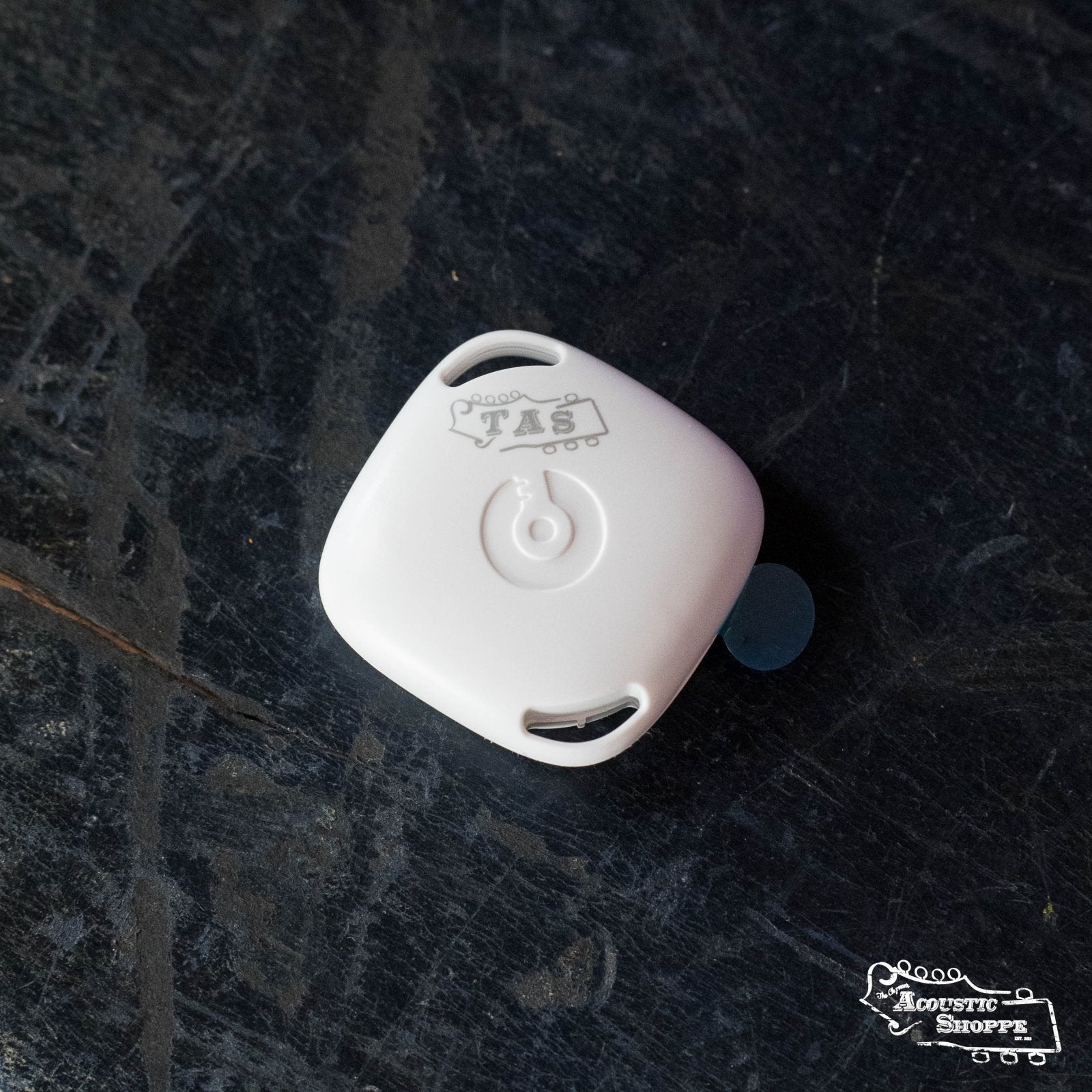 A compact, square white AcoustiCare Bluetooth Humidity and Temperature Tracker by The Acoustic Shoppe rests on a dark textured surface. It features rounded edges, two openings, a center button, and The Acoustic Shoppe logo in the bottom right.