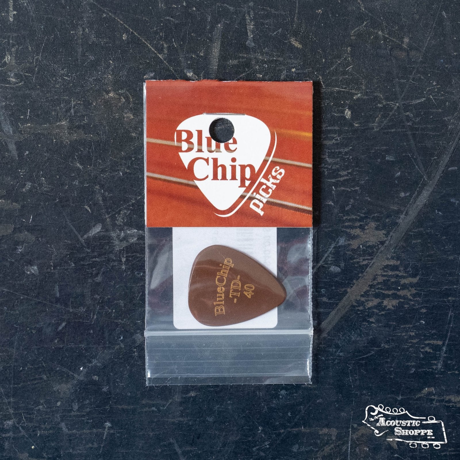 A BlueChip Picks (TD40) by BlueChip, in red and white packaging, rests on a dark scratched surface. The brown teardrop pick with gold lettering is visible inside, with a white logo at the bottom right of the package.