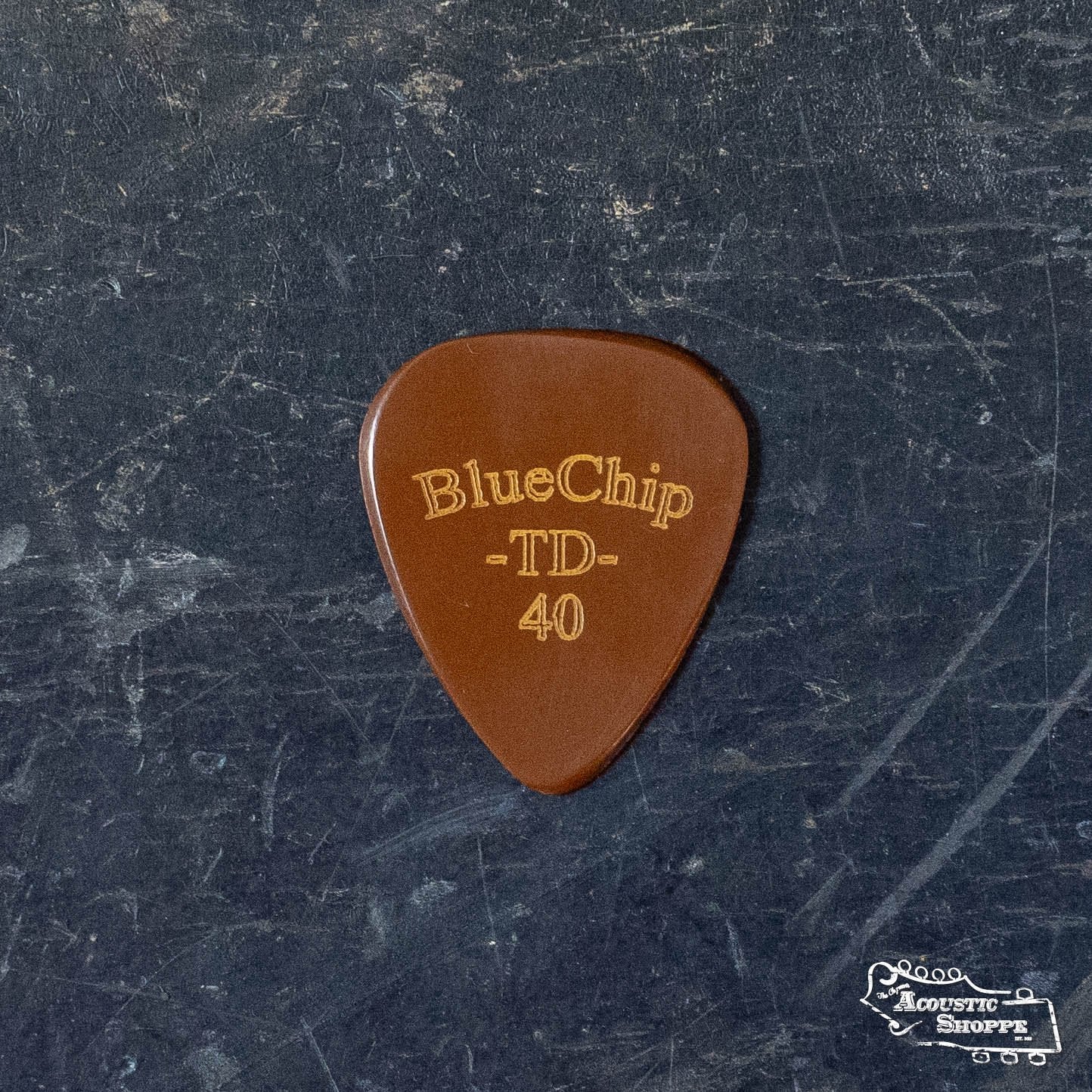 A BlueChip Picks (TD40) guitar pick with rounded edges rests on a dark, worn surface, sporting a small The Acoustic Shoppe logo in the bottom right corner—a premium accessory from BlueChip.