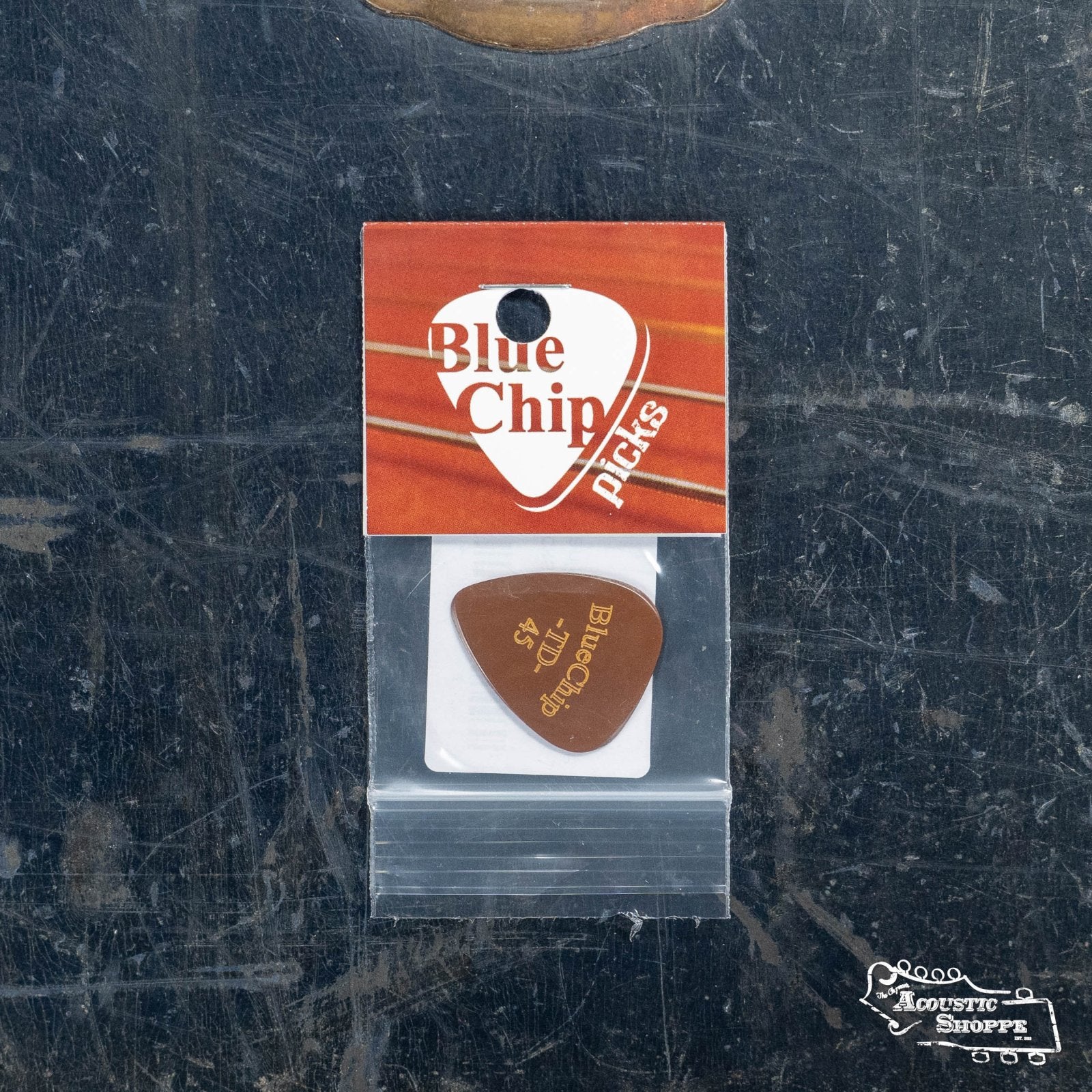 A BlueChip Picks (TD45) teardrop guitar pick by BlueChip comes in a small plastic bag attached to an orange-accented BlueChip card, displayed on a dark, scratched metal surface—ideal for acoustic music enthusiasts.