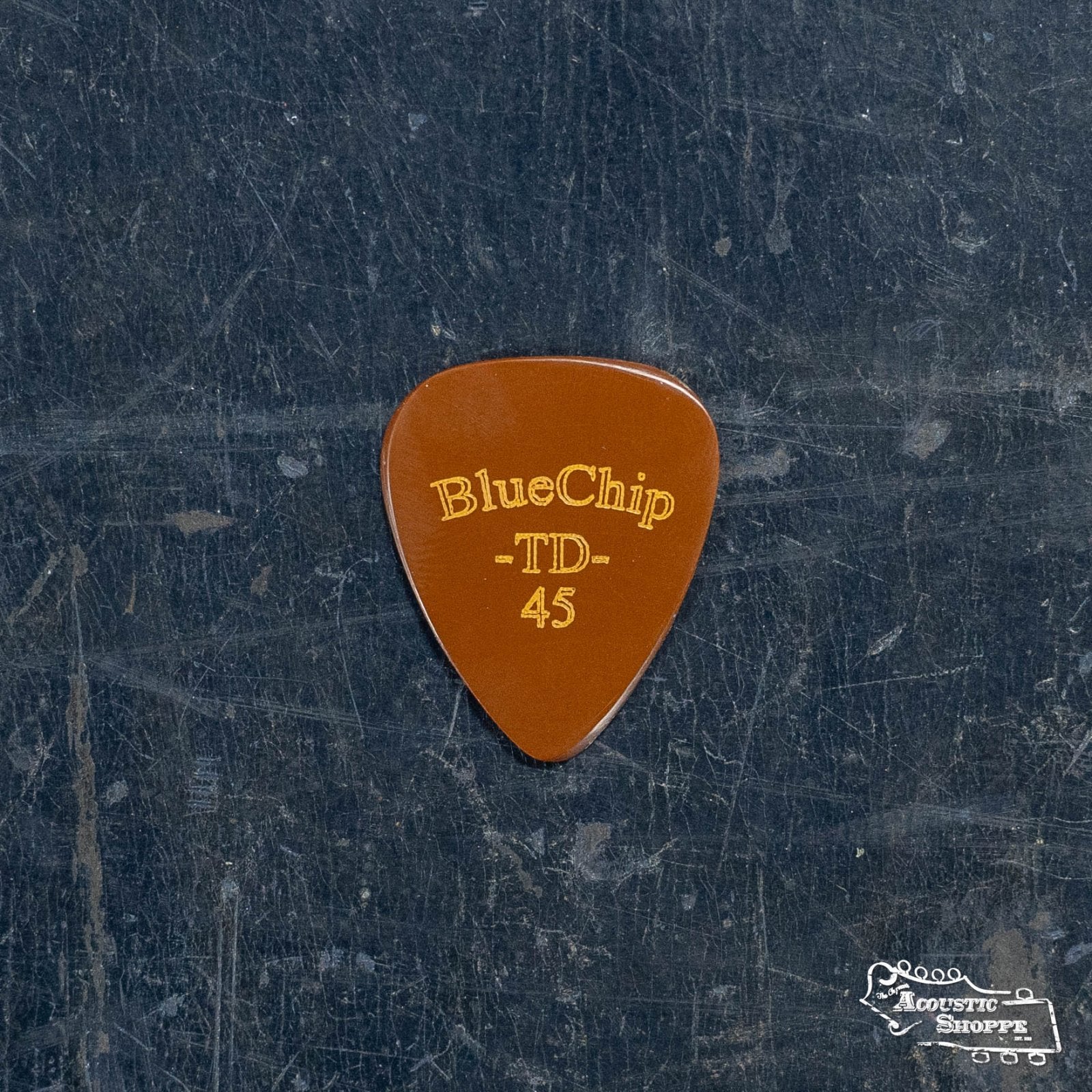 A BlueChip Picks (TD45) guitar pick with gold lettering rests on a worn, dark surface. A small white Acoustic Shoppe logo in the lower right corner highlights quality BlueChip acoustic accessories.