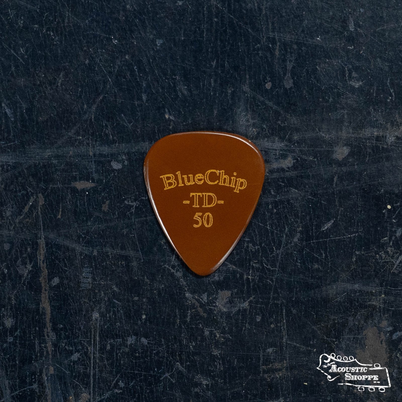 A BlueChip Picks (TD50) brown teardrop guitar pick with gold BlueChip text rests on a dark, scratched surface. The Acoustic Shoppe logo appears in the bottom right, highlighting premium acoustic accessories.