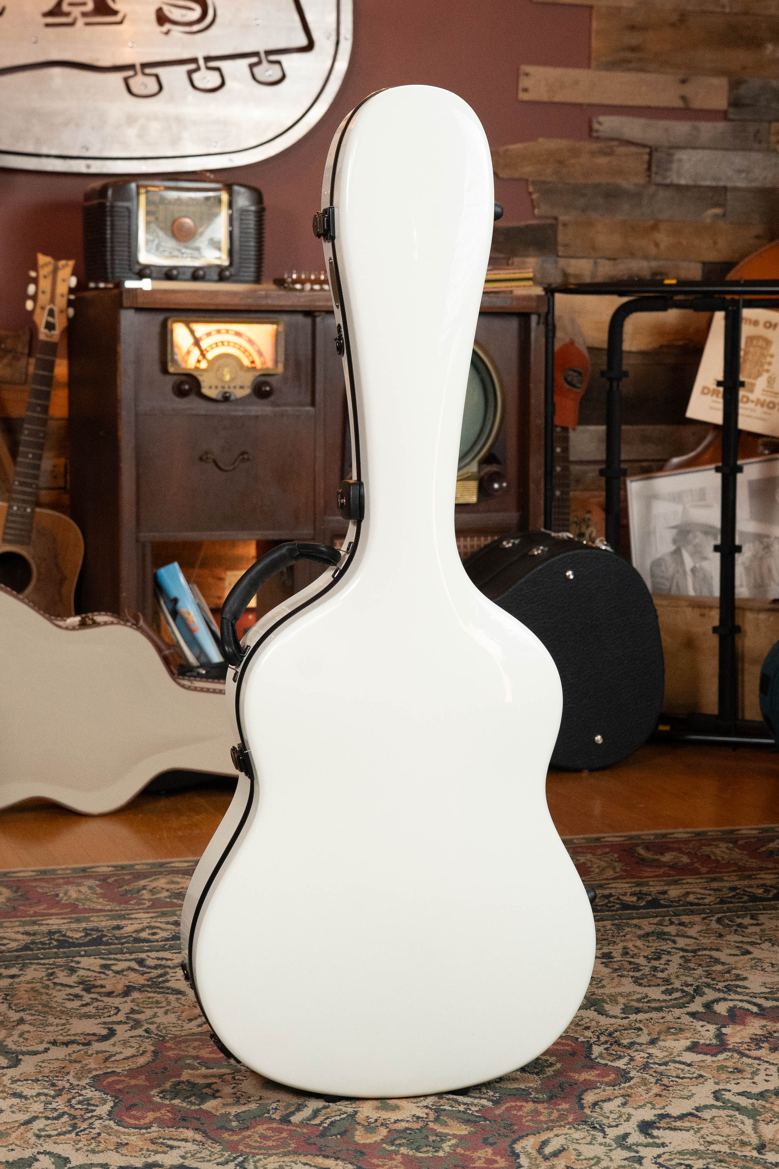 The TKL Voyager Universal Pearl Glacier/Blue Carbon Fiber Dreadnought Case stands upright in a cozy room with vintage decor, surrounded by an old radio, wooden cabinets, other guitar cases, and a patterned rug.