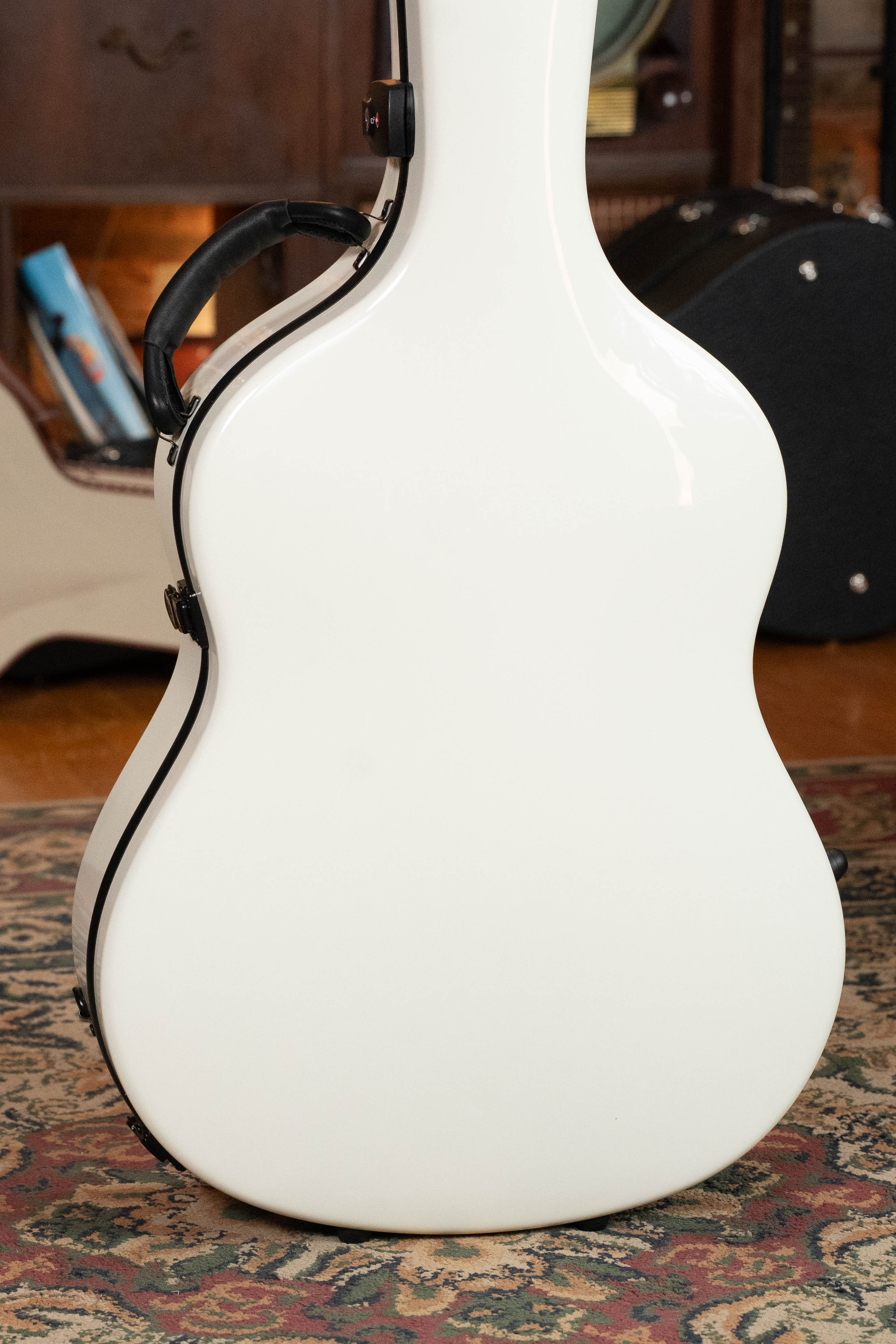 A TKL Voyager Universal Pearl Glacier/Blue Carbon Fiber Dreadnought Case stands upright on a patterned rug, its white lightweight design with black trim in focus, while a bookshelf and another guitar case blur in the background.