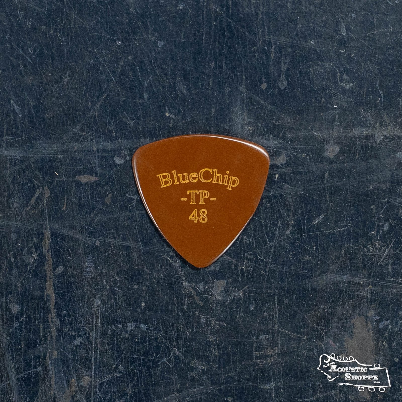 A brown BlueChip Picks (TP48) triangular pick, featuring gold lettering, rests on a dark textured surface—a top-tier acoustic guitar accessory from BlueChip.