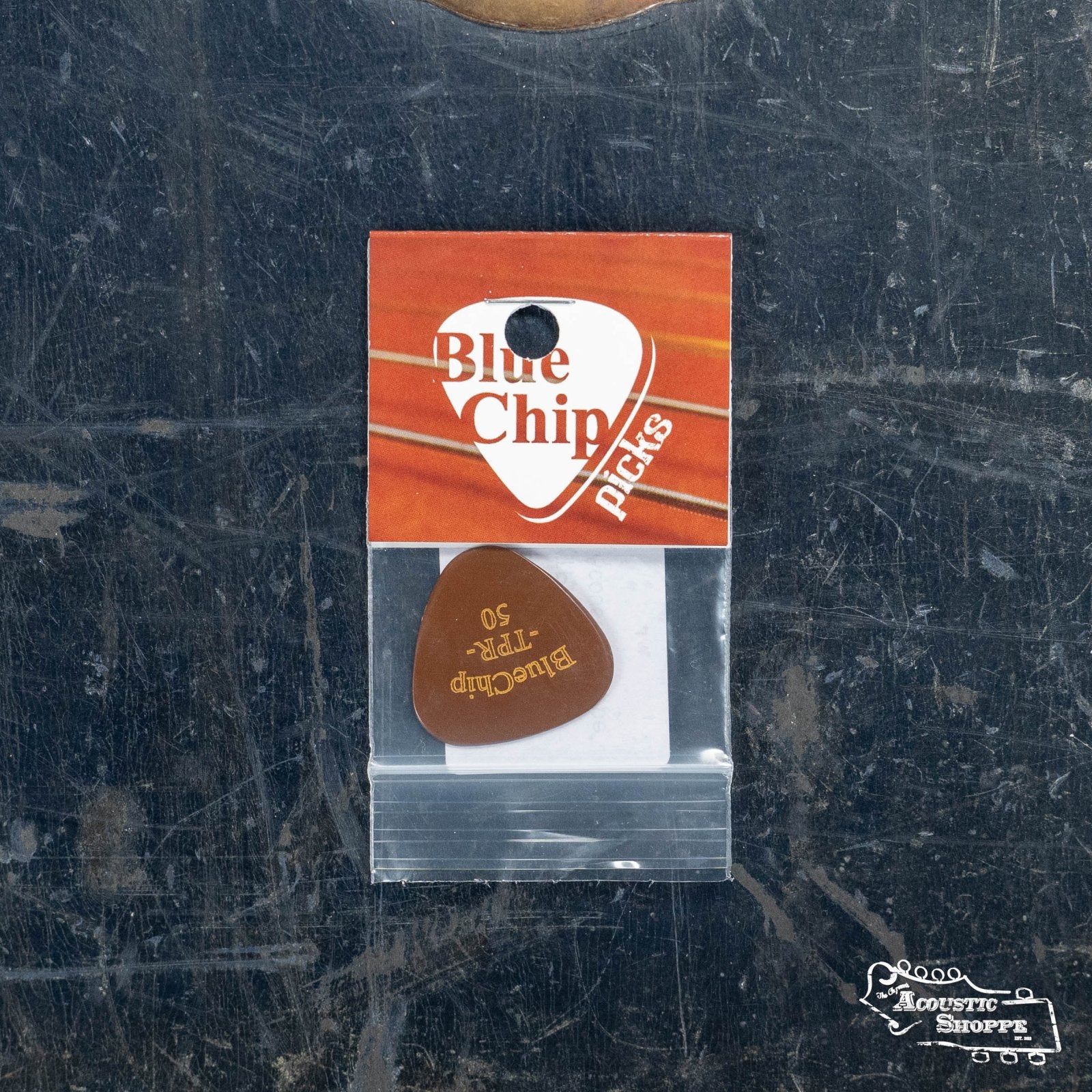 A BlueChip Picks (TPR50) guitar pick from BlueChip, in a plastic bag with an orange and white label, sits on a dark, scratched surface. The Acoustic Shoppe logo is at the bottom right—ideal for acoustic guitar players.