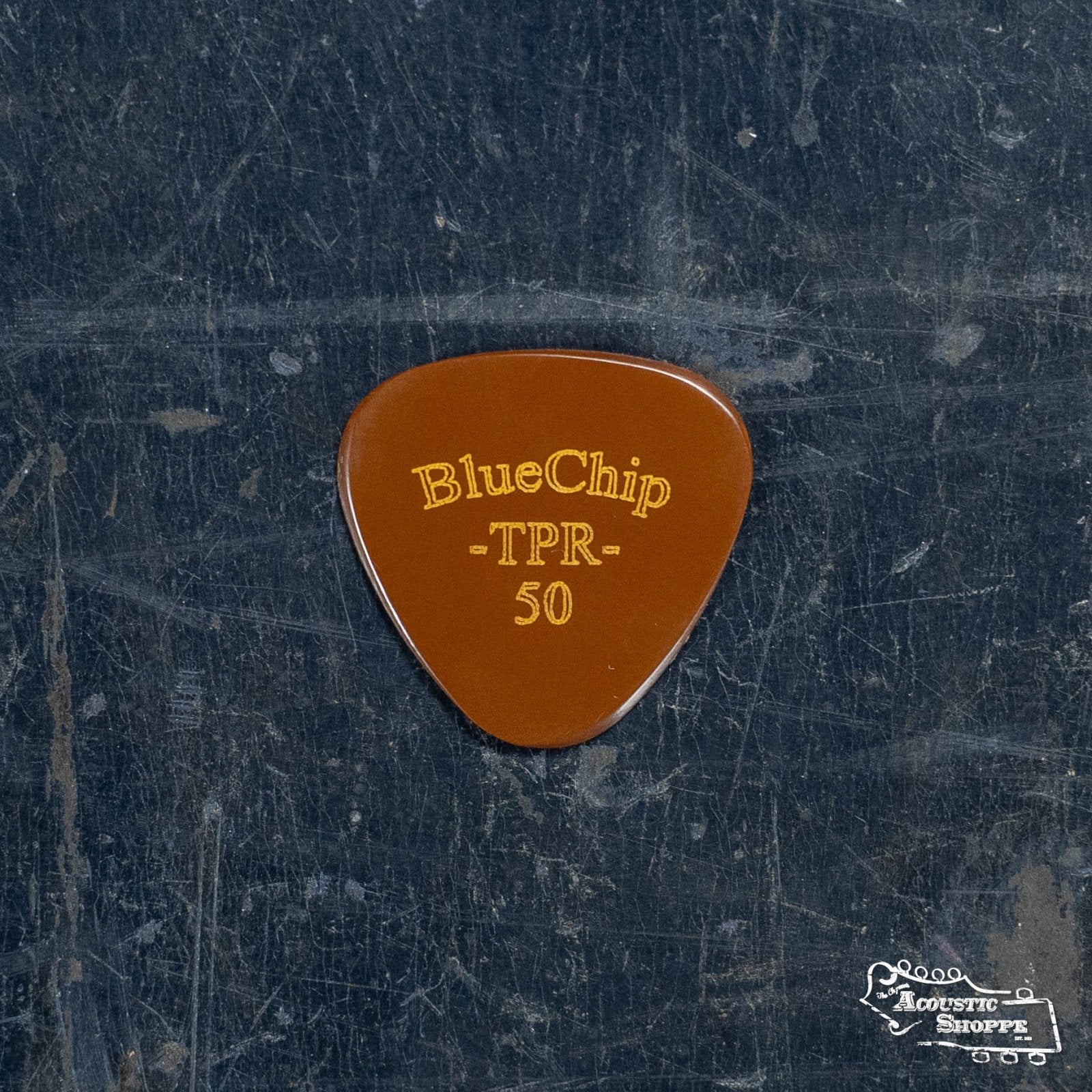 A BlueChip Picks (TPR50) guitar pick in brown, popular with acoustic fans, sits on a scratched dark surface featuring the white Acoustic Shoppe logo in the bottom right corner.