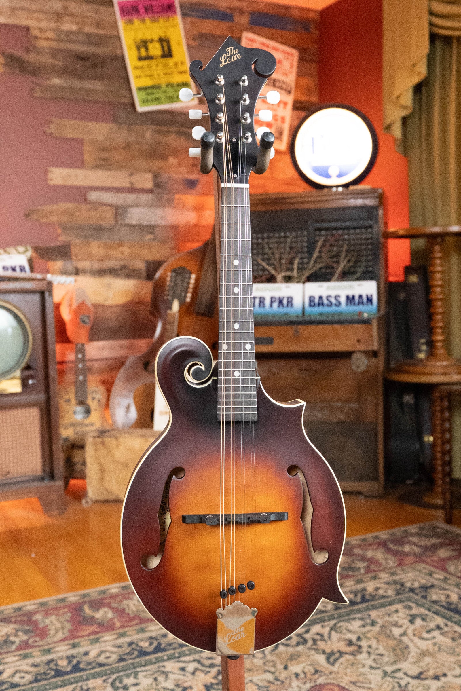 A The Loar LM-310F-BRB Honey Creek F-Style Brownburst Mandolin #0069 stands on a patterned rug in a cozy, vintage-style room filled with musical instruments, posters, and antique audio equipment.