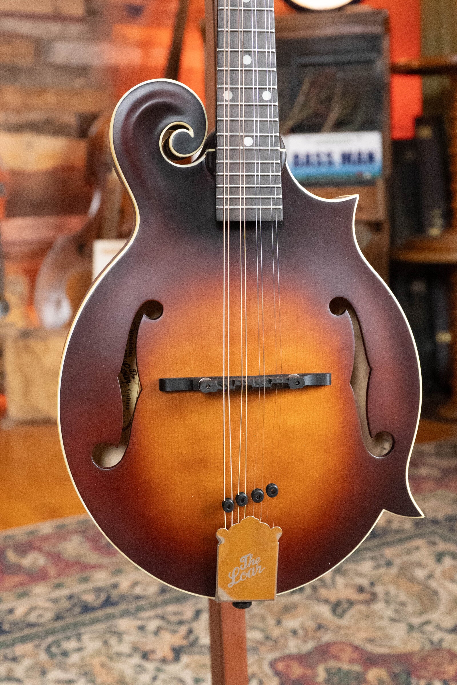 A close-up of The Loar LM-310F-BRB Honey Creek F-Style Brownburst Mandolin #0069, featuring a hand-carved spruce top and The Loar tailpiece, set amid musical equipment and a patterned rug.