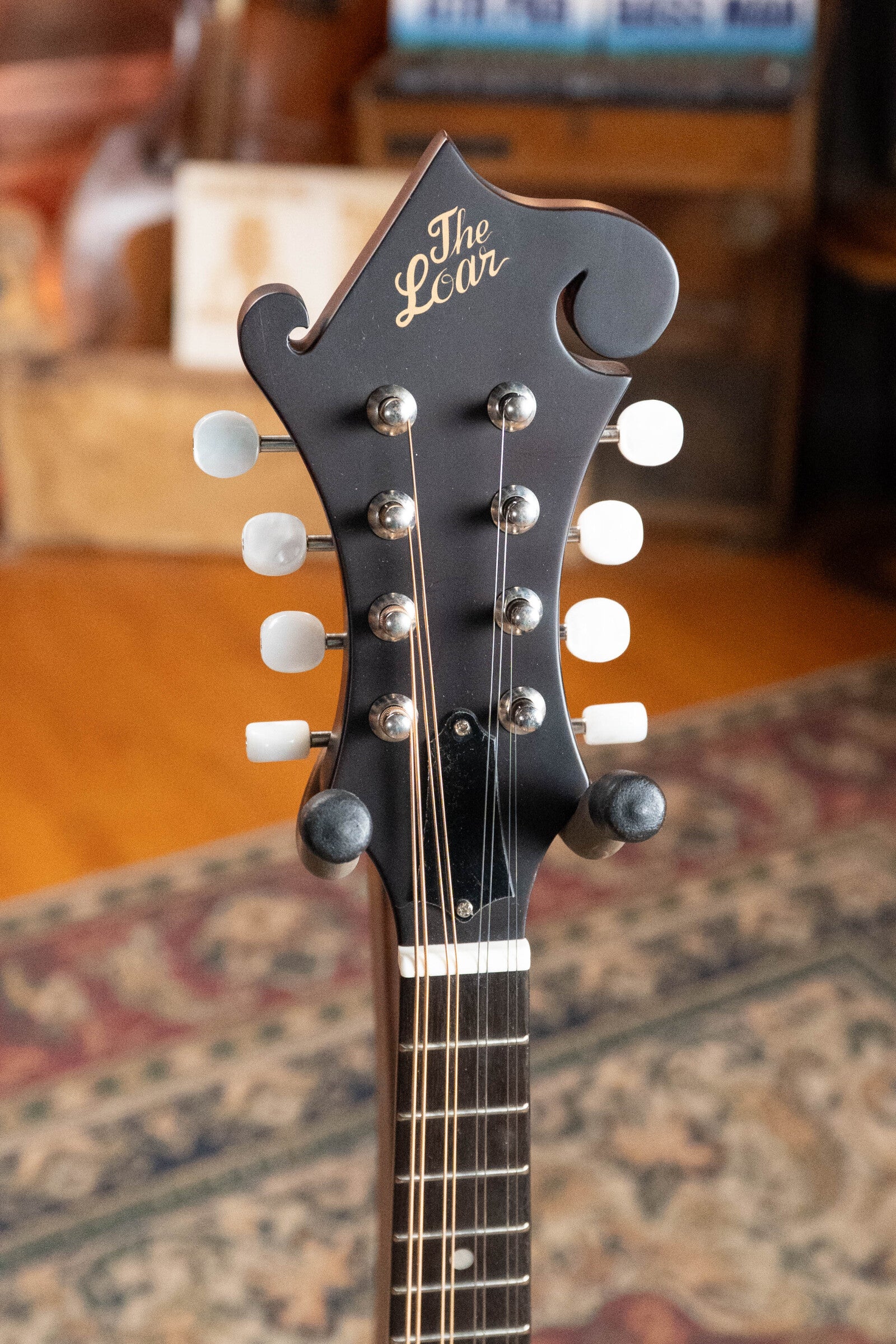 A close-up of The Loar LM-310F-BRB Honey Creek F-Style Brownburst Mandolin #0069 headstock, showing the script logo, white tuning pegs, and metal posts against a blurred indoor background with a patterned rug.