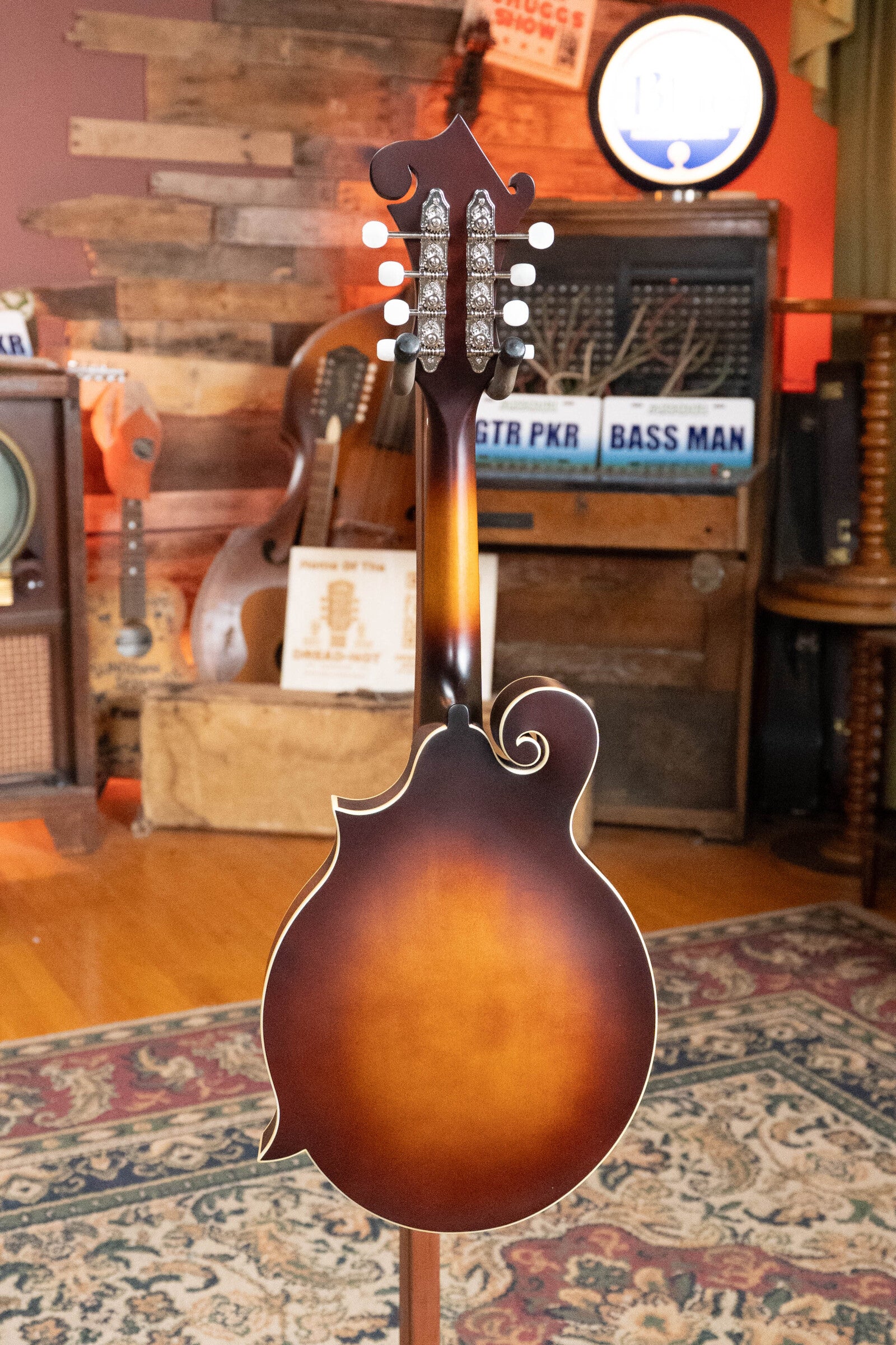 The Loar LM-310F-BRB Honey Creek F-Style Brownburst Mandolin #0069 by The Loar, with a hand-carved spruce top and decorative scroll, is displayed on a patterned rug in a cozy vintage music room filled with instruments and wooden decor.