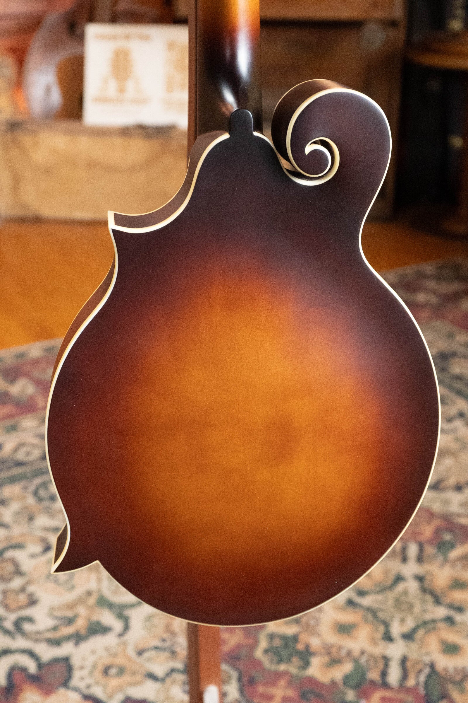 A close-up shows the back of The Loar LM-310F-BRB Honey Creek F-Style Brownburst Mandolin #0069, featuring decorative binding and scroll design, resting on a patterned rug in a warmly lit room. Brand: The Loar.