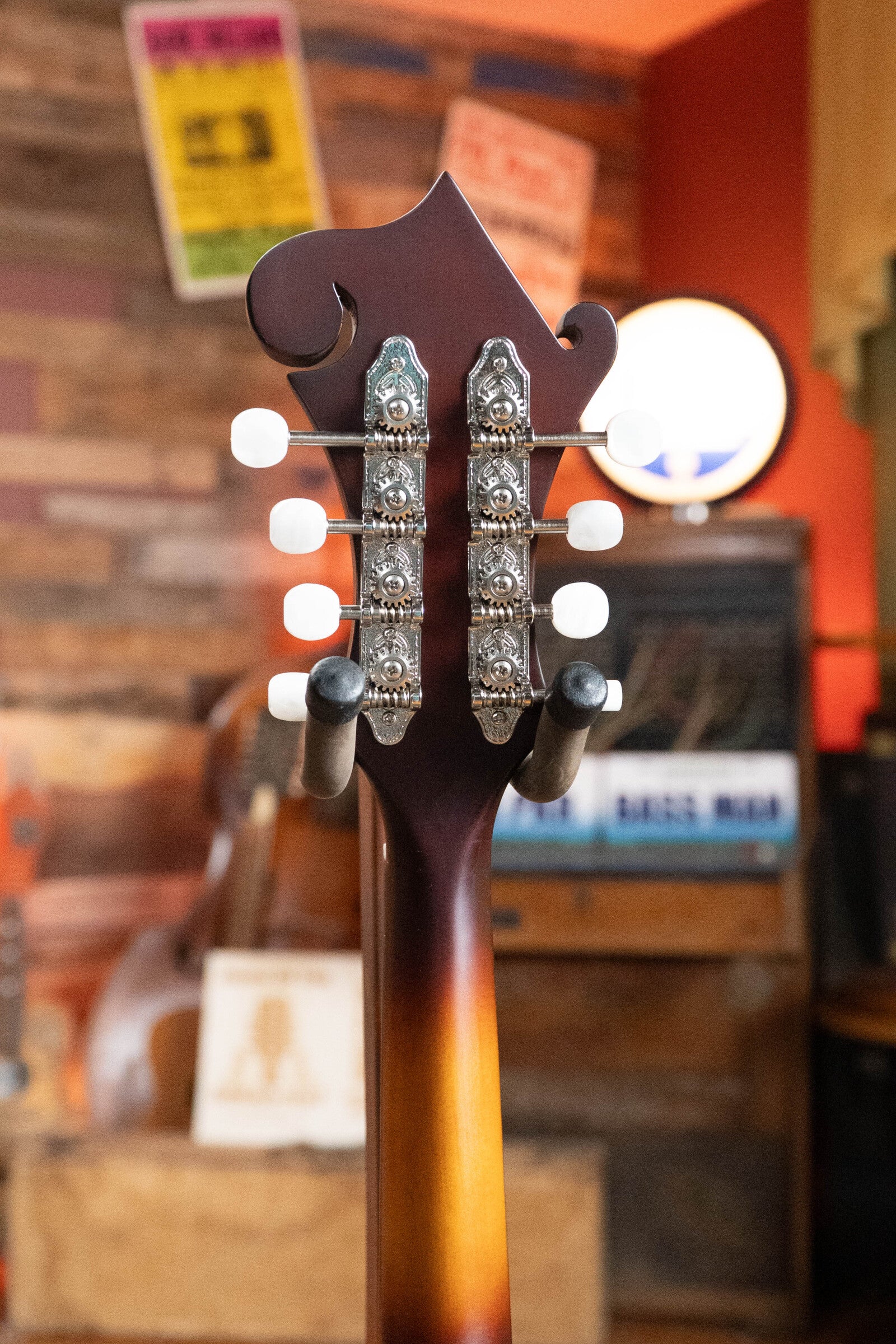 Close-up of the back of The Loar LM-310F-BRB Honey Creek F-Style Brownburst Mandolin #0069 headstock, showing tuning pegs and ornate metal plates; rustic blurred background with wood paneling, posters, and musical equipment.
