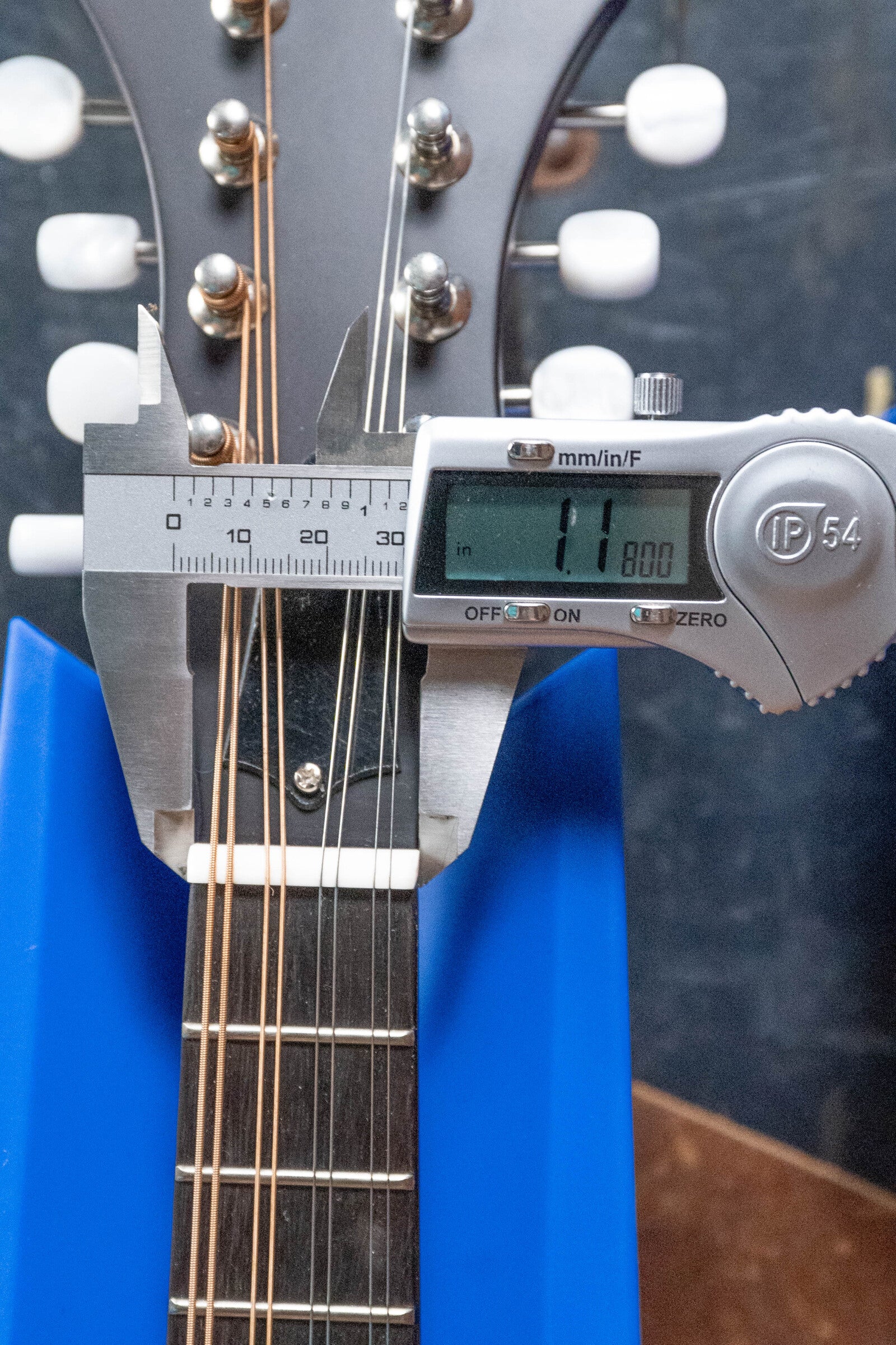 A digital caliper shows the neck width at the nut of The Loar LM-310F-BRB Honey Creek F-Style Brownburst Mandolin #0069 as 11.80mm. The Loar mandolin sits in a blue stand, with strings and tuning pegs visible.