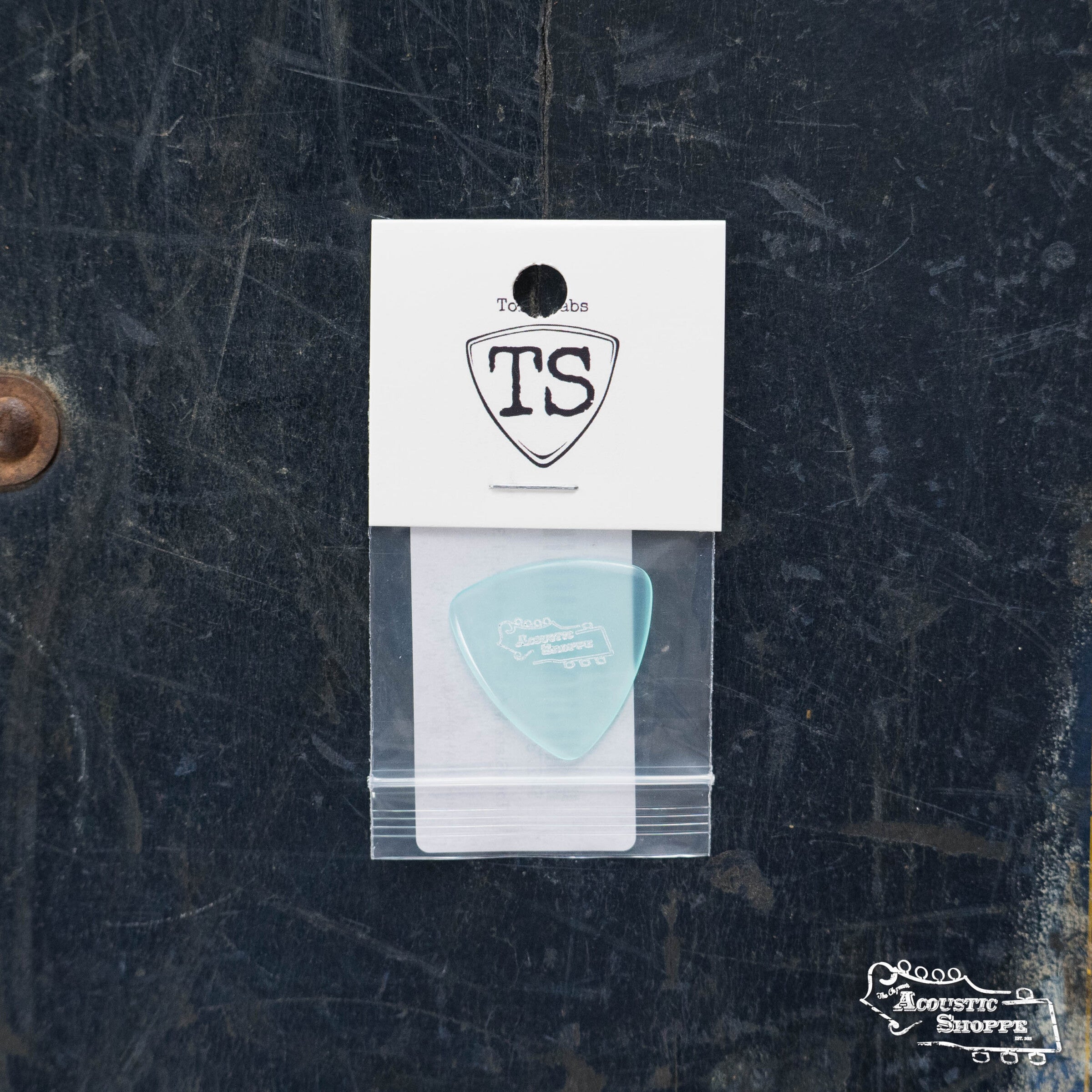 A light blue ToneSlabs Mini Tri Guitar Pick - 1.3mm, packaged in a clear plastic bag with a white TS card and guitar pick graphic, is shown on a dark, worn surface.