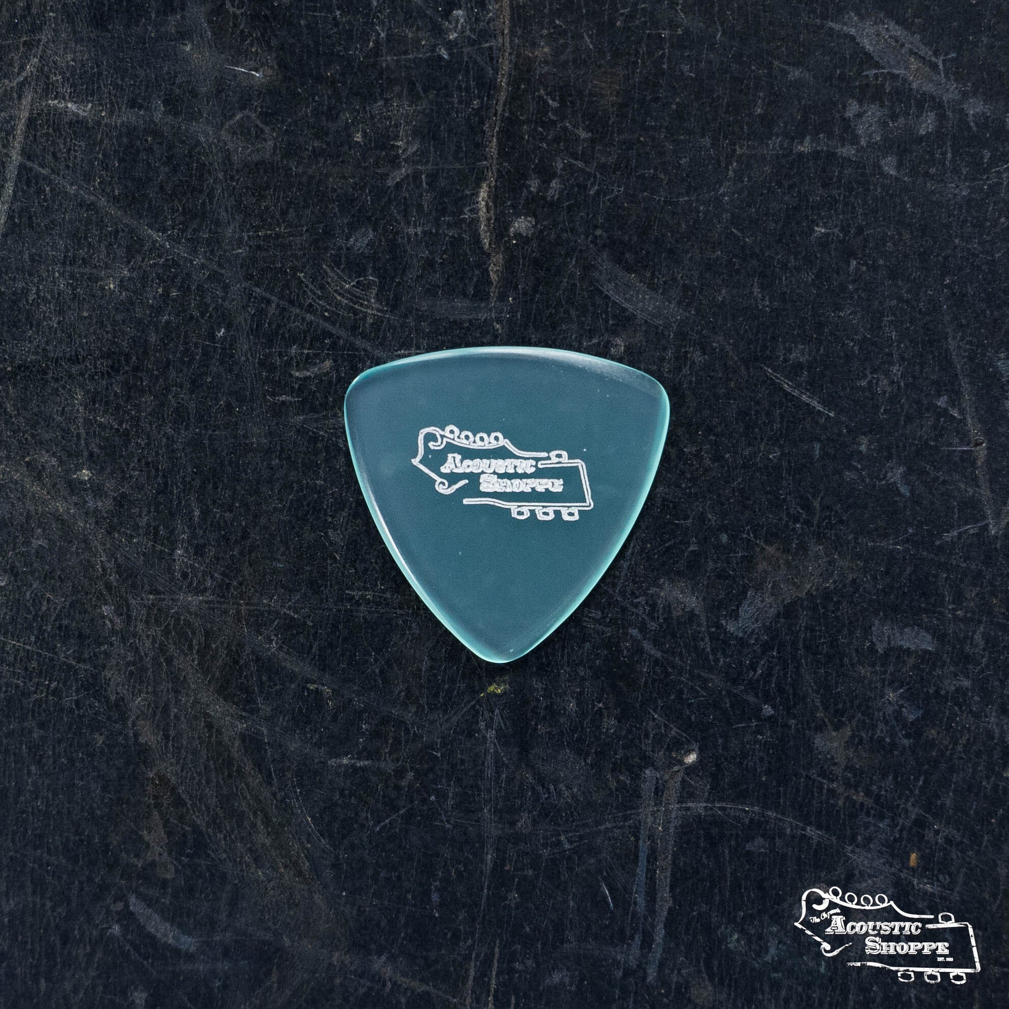 A translucent blue ToneSlabs Mini Tri Guitar Pick - 1.3mm with The Acoustic Shoppe logo rests on a dark textured surface, with the same logo visible in the lower right corner.