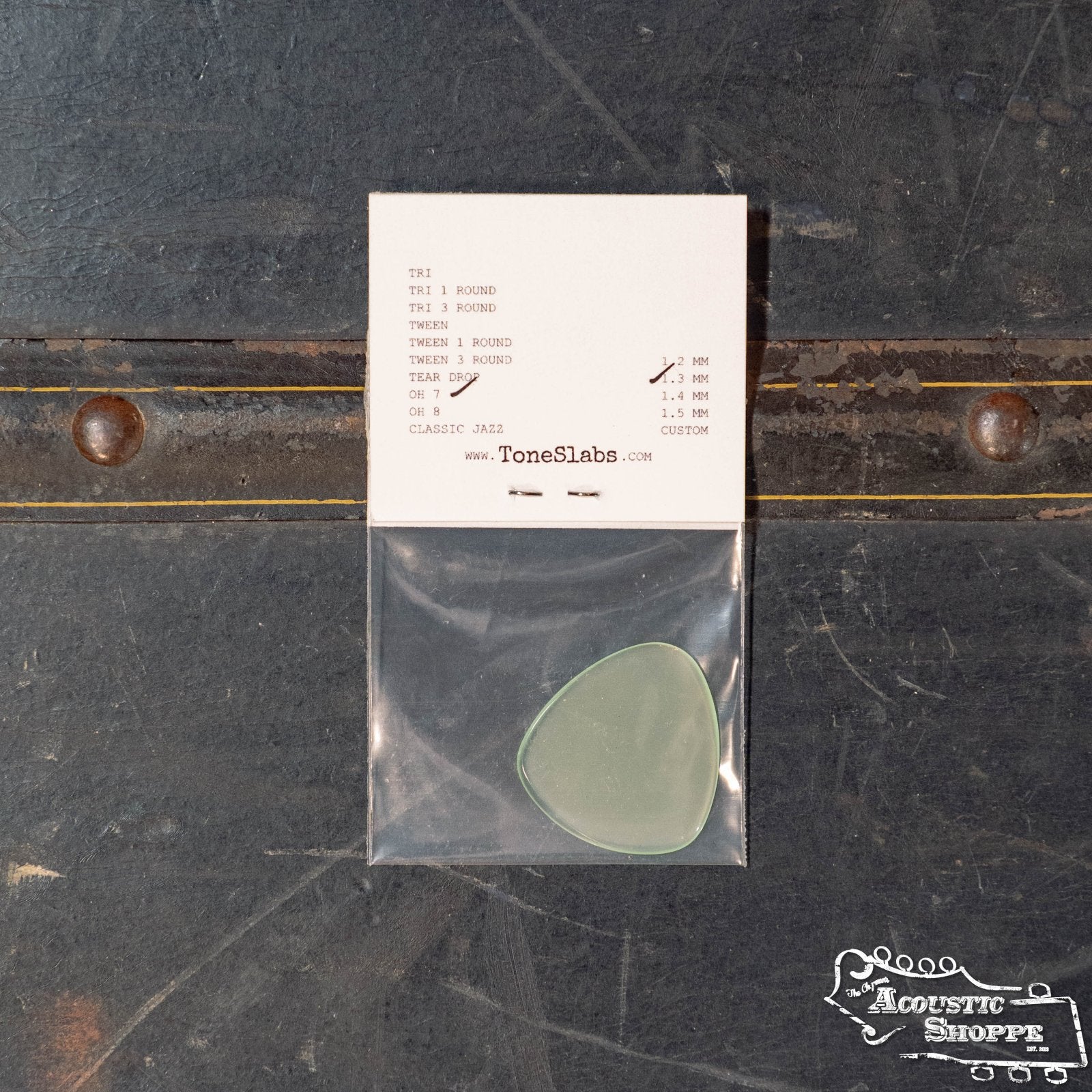 A translucent green ToneSlabs Oh Seven Rounded Jazz Guitar Pick (1.3 mm) in a plastic bag sits on a dark surface near a label card and metal rivets. The ToneSlabs logo in the corner highlights its premium quality.