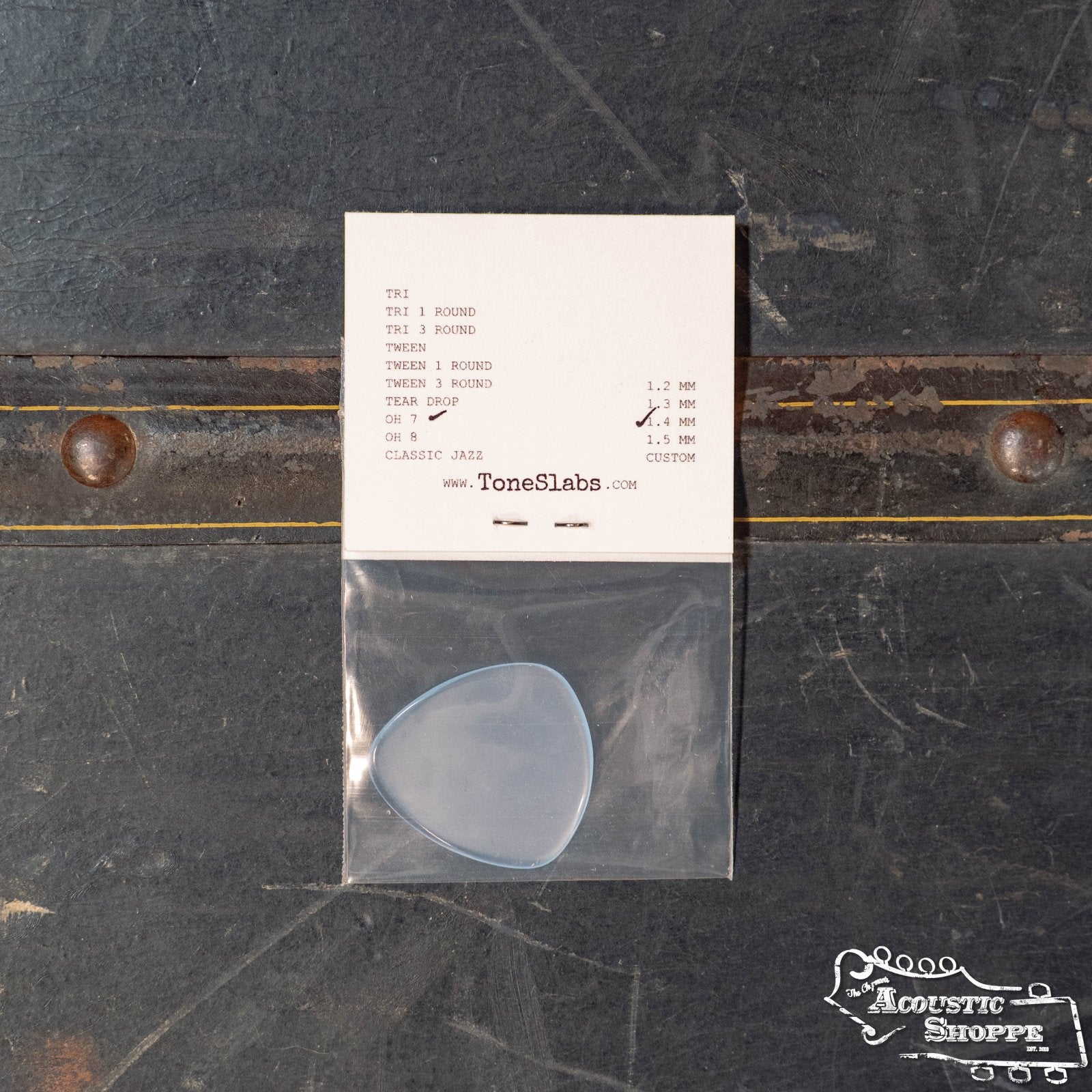 The ToneSlabs Oh Seven Rounded Jazz Guitar Pick (1.4 mm) by ToneSlabs comes in a small plastic bag attached to a card showing other models, including tortoise shell replacements and tone slab styles, on a dark surface with The Acoustic Shoppe logo.