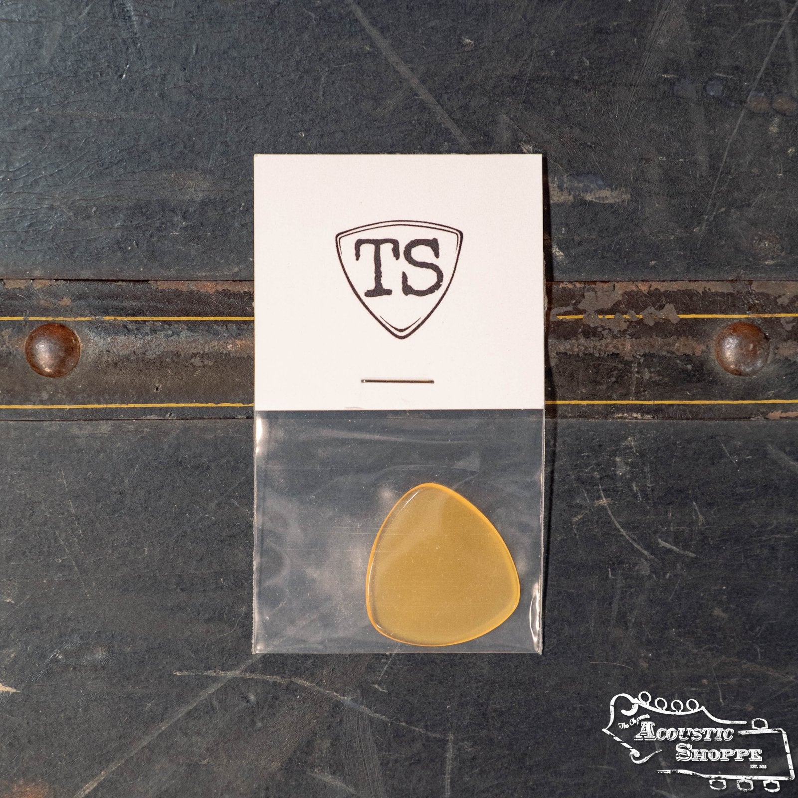 A ToneSlabs Oh Seven Rounded Jazz Guitar Pick (1.5 mm, tan tortoise shell) in a small clear bag sits on a dark textured surface. Above it is a shield logo card with “TS,” and The Acoustic Shoppe logo is in the bottom right corner.