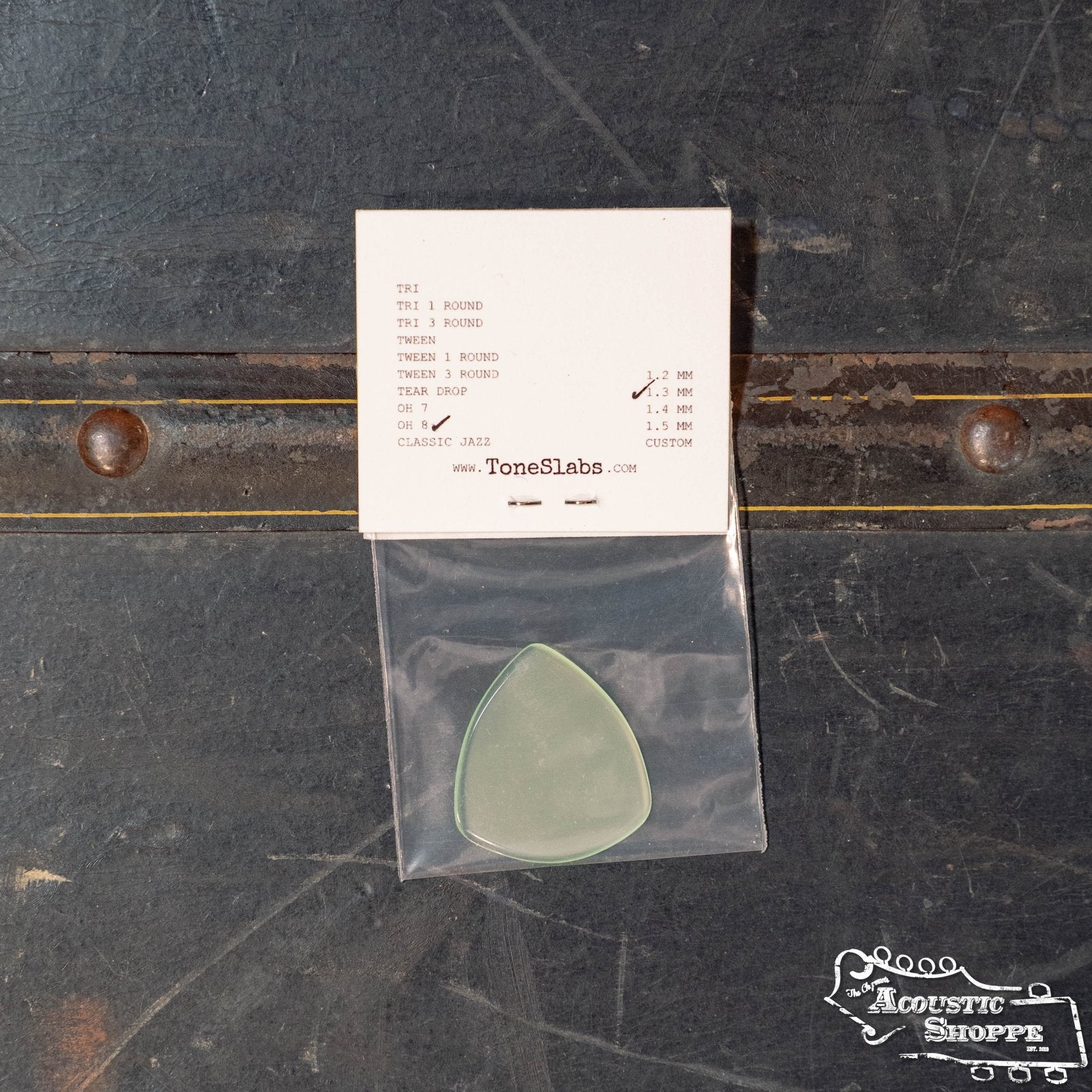A green ToneSlabs Oh Eight Pointed Jazz Guitar Pick (1.3 mm) rests in a clear pouch on a dark surface, with a white ToneSlabs card listing pick options above it. Both items feature the ToneSlabs brand and Acoustic Shoppe logo.