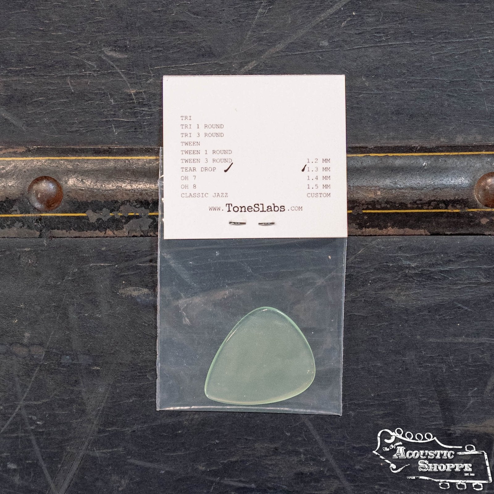 The ToneSlabs TAS Custom Tear Drop Guitar Pick, 1.3 mm (Exclusive Color), sits in a clear bag on a dark surface with specs on a white slip behind it. The Acoustic Shoppe logo adds appeal for acoustic fans seeking premium picks.