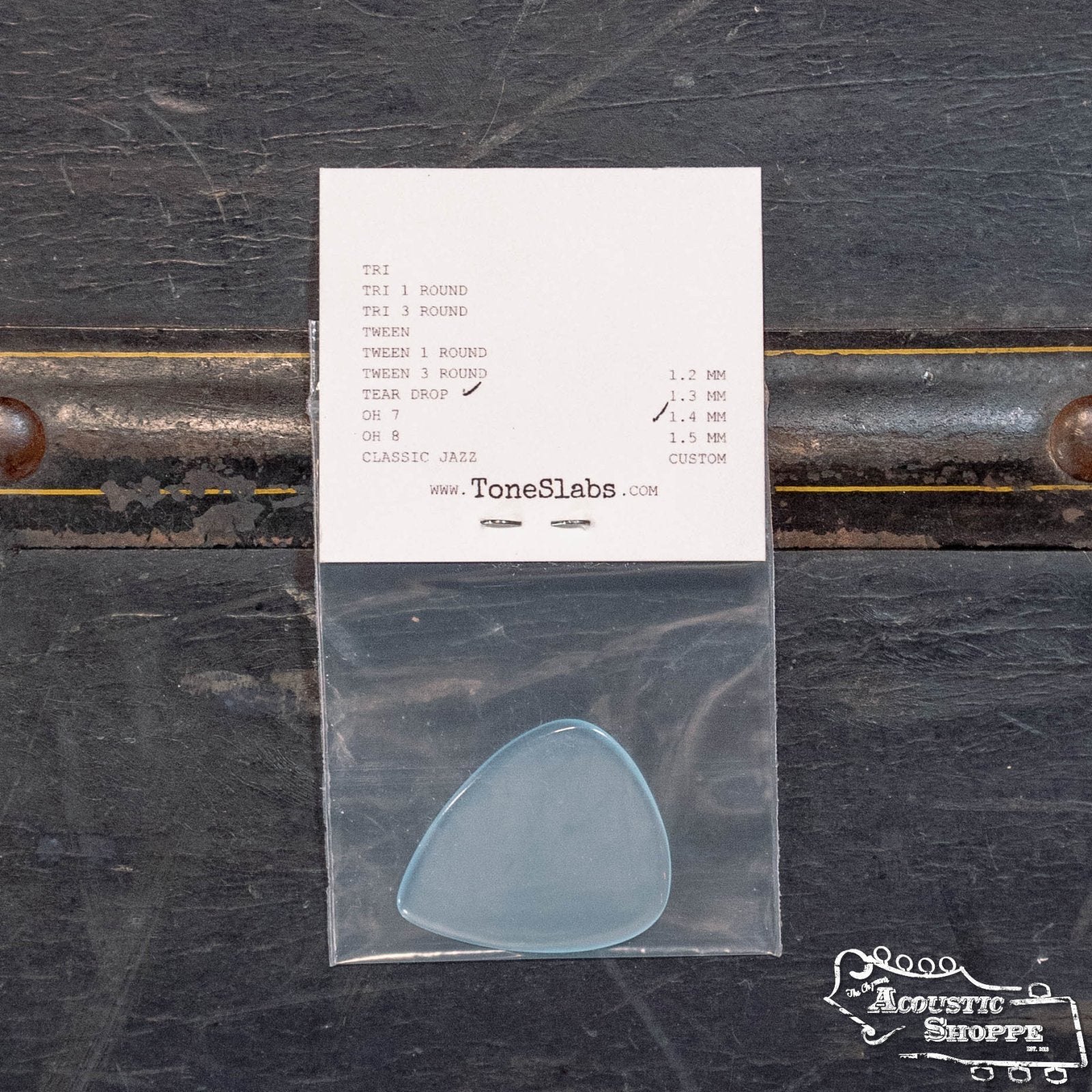 A ToneSlabs Tear Drop Guitar Pick (1.4 mm) comes in a blue teardrop shape, packaged in a plastic bag on a white card displaying pick options. Placed on a dark textured surface, it features the Acoustic Shoppe logo and ToneSlabs branding.