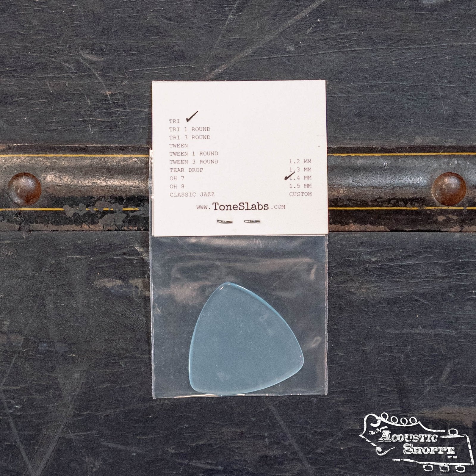 A ToneSlabs XL Tri Corner Guitar Pick (1.4 mm) in blue, packaged in a plastic bag, lies with a pick shape and thickness info card on a dark, worn surface with metal rivets. The Acoustic Shoppe logo is at the lower right corner.