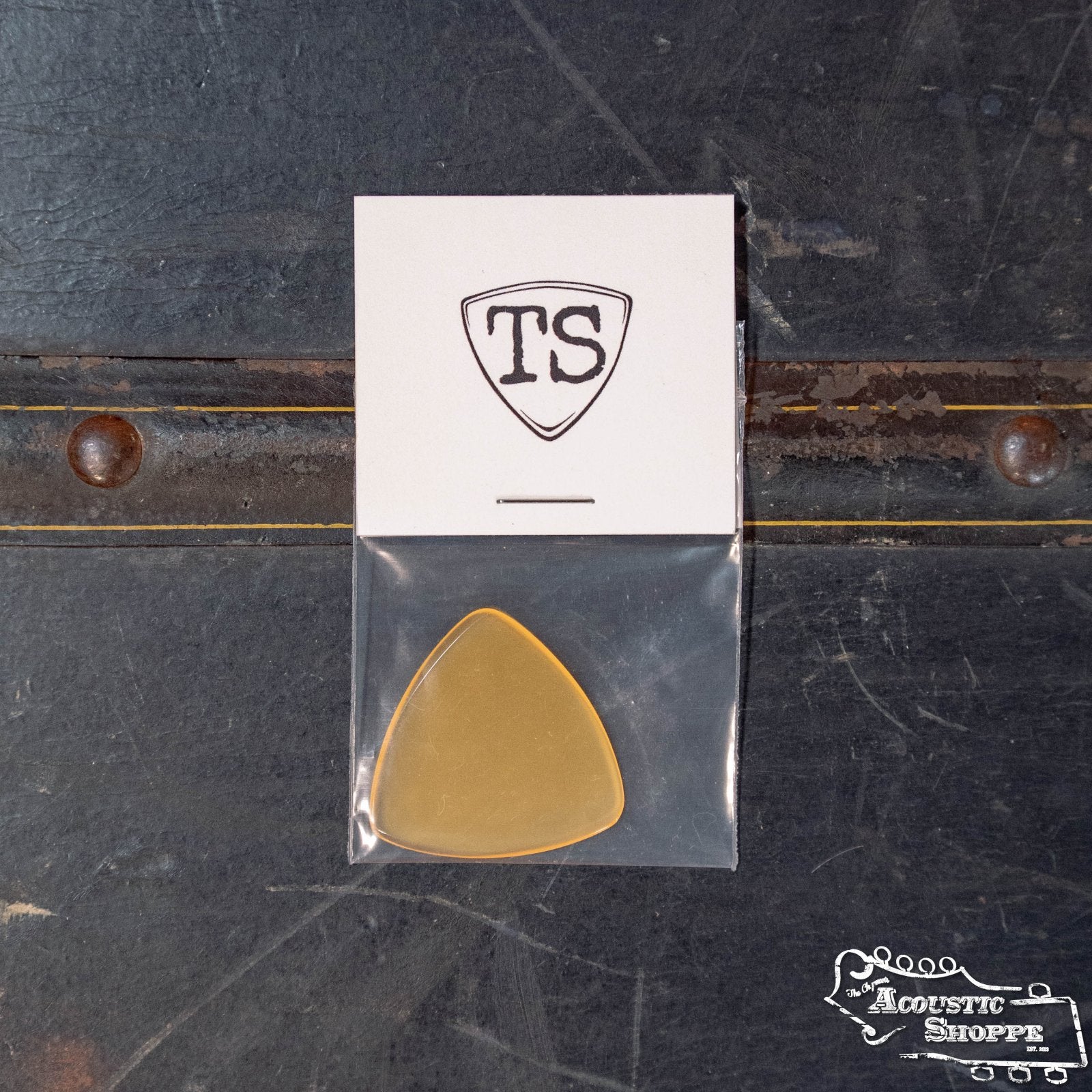 A ToneSlabs XL Tri Corner Guitar Pick (1.5 mm) in gold rests in a clear sleeve on a dark surface. Above is a white card with the TS shield design, and The Acoustic Shoppe logo is on the bottom right.