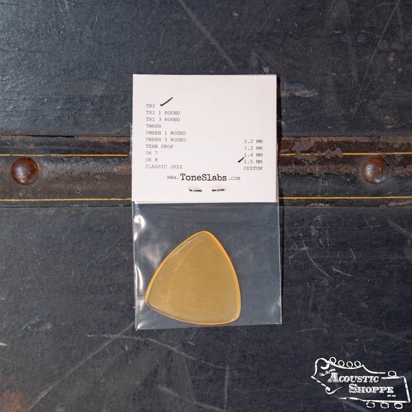 A ToneSlabs XL Tri Corner Guitar Pick (1.5 mm) rests in a clear plastic sleeve below a white card listing pick types and pricing, all on a dark wooden surface. The ToneSlabs brand is featured in the lower right corner.